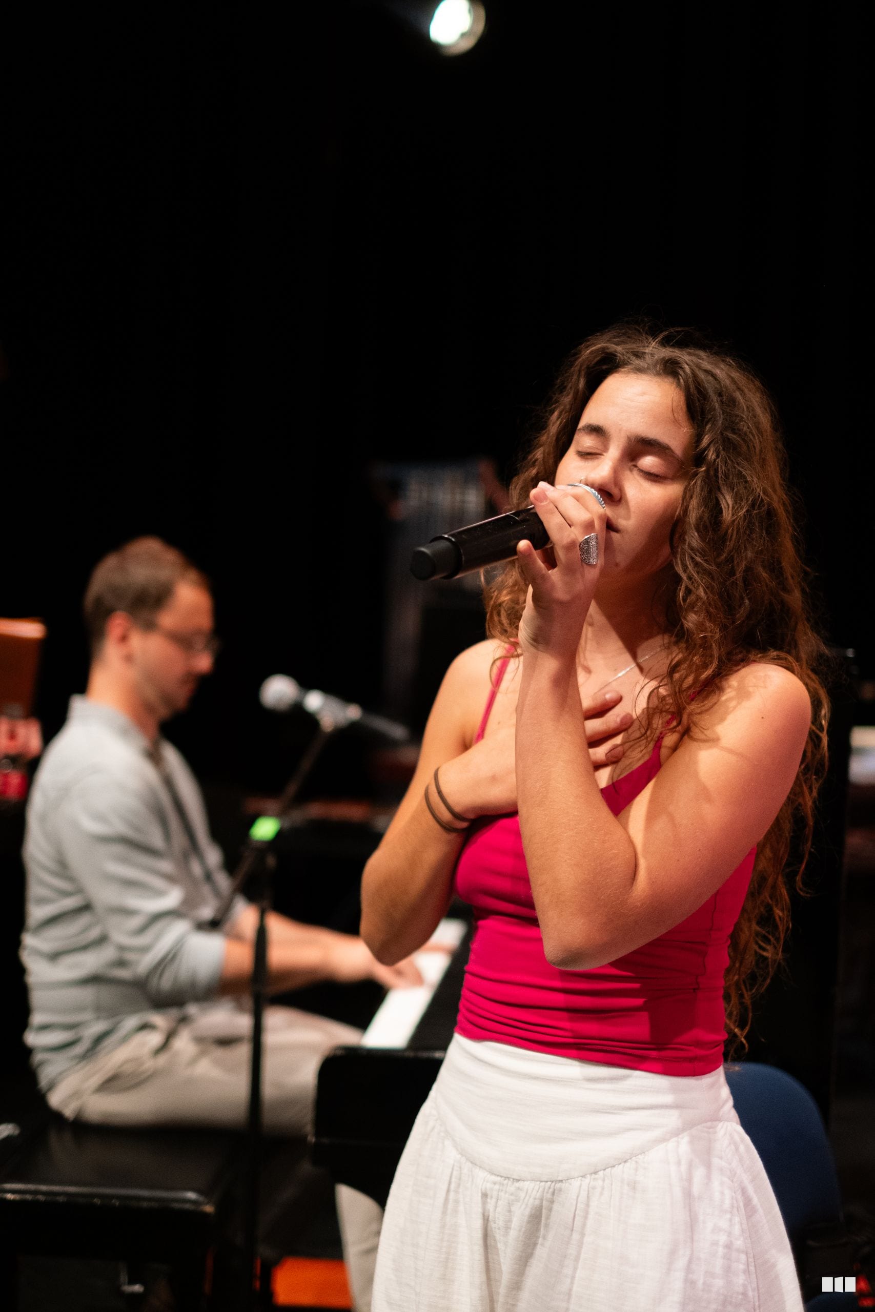 A female soloist performs with a microphone while a pianist accompanies her on stage.