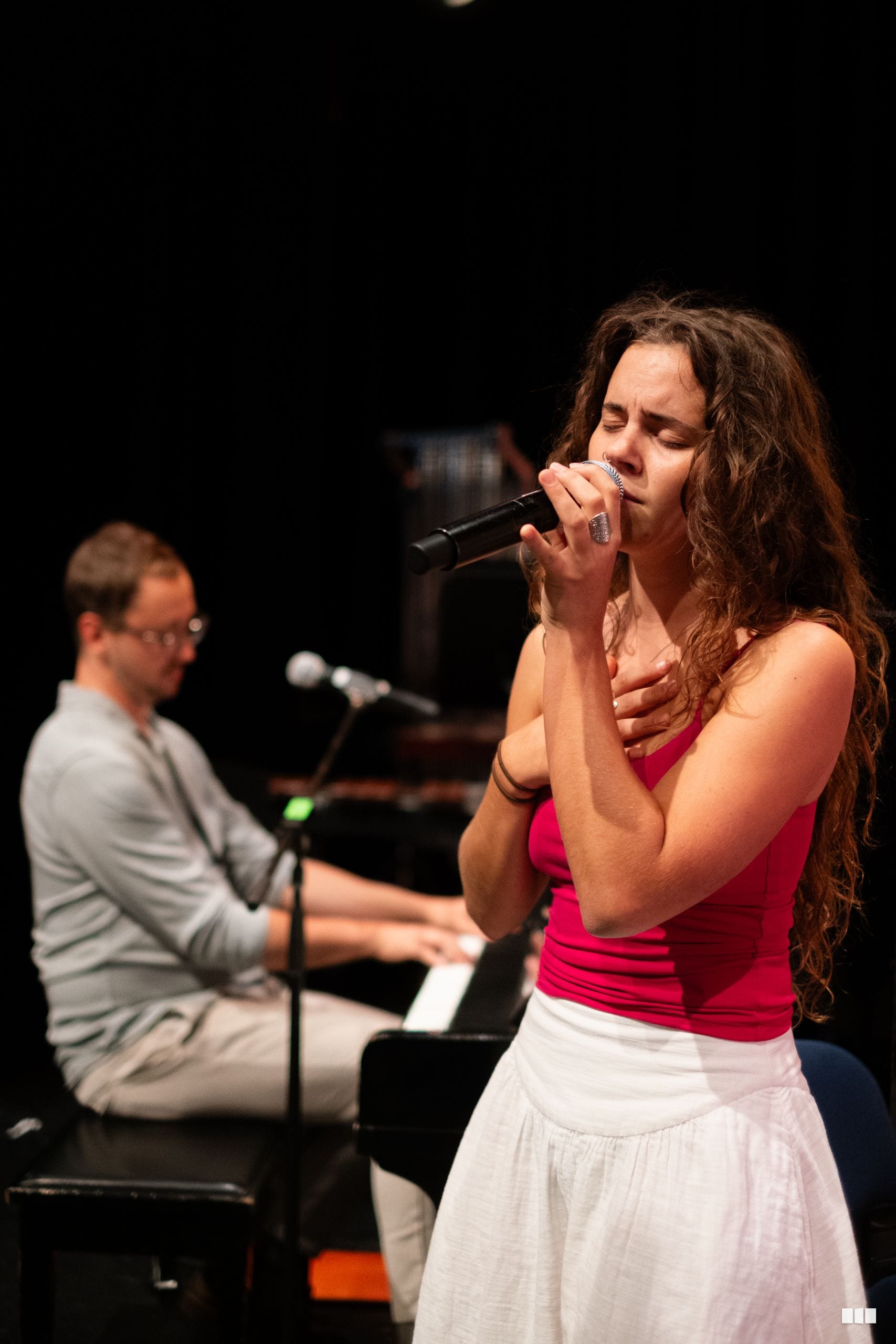 A female soloist performs with a microphone while a pianist accompanies her on stage.