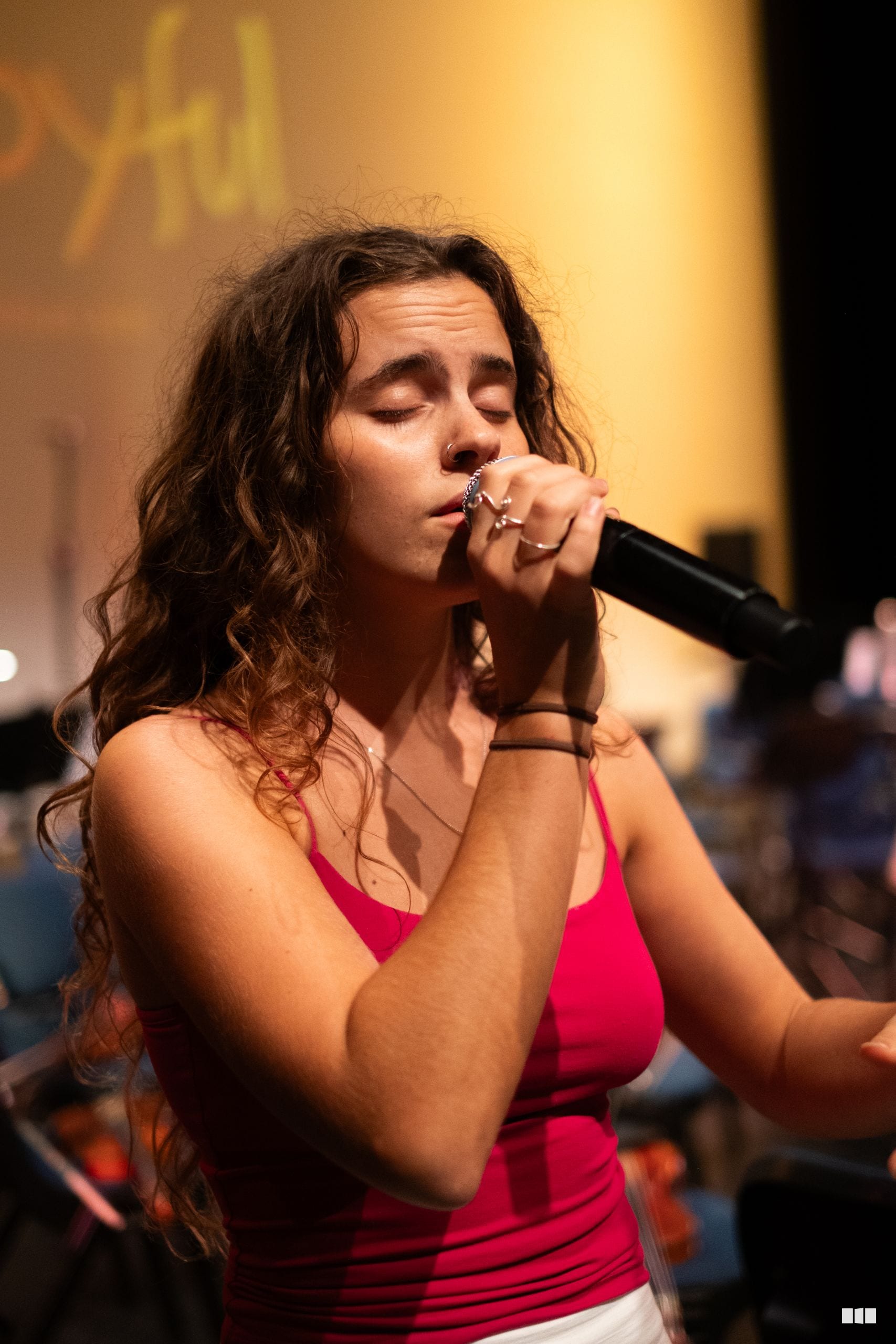 A female soloist performs passionately with a microphone against a warm amber-lit stage background.