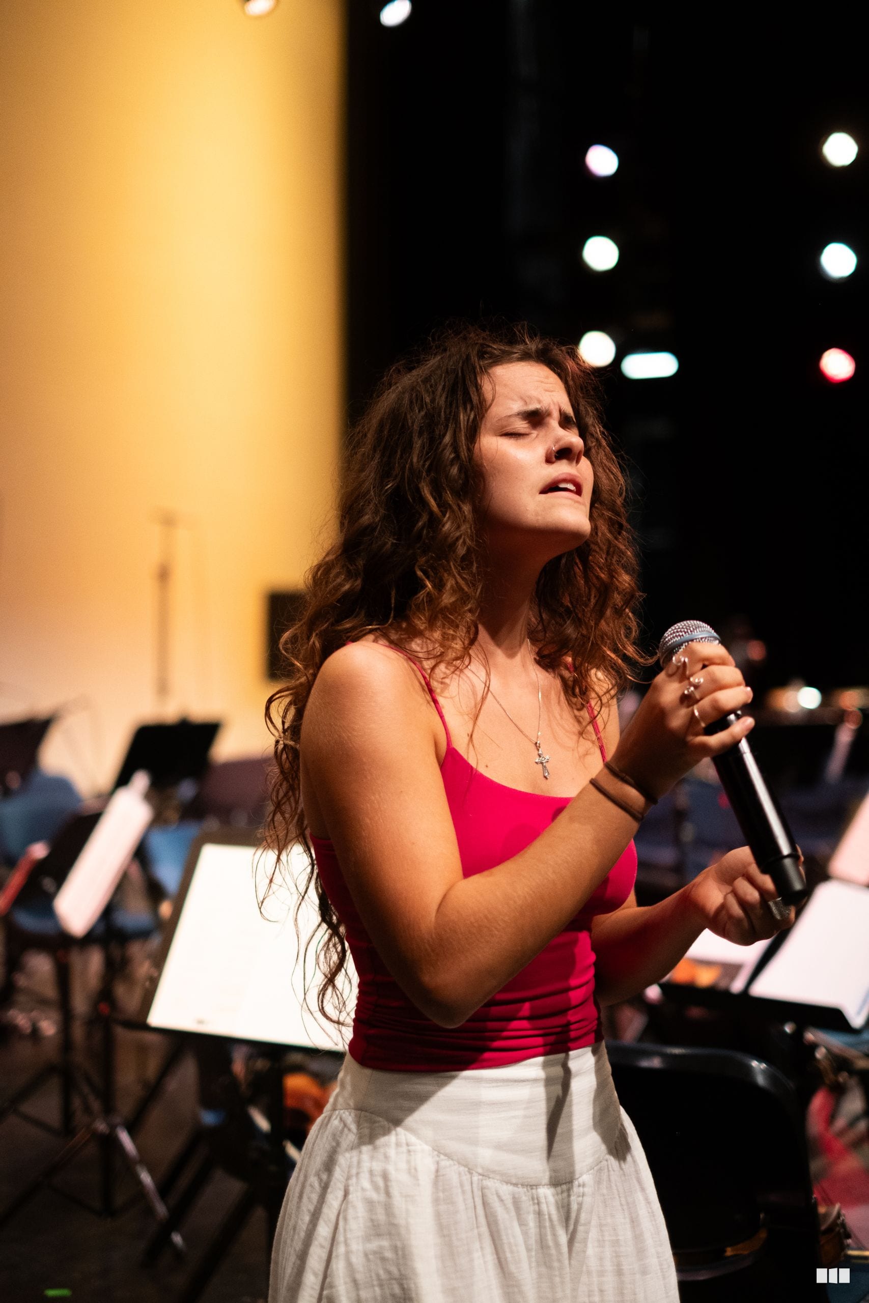 A young female soloist in a red top performs with a microphone in front of an orchestra during an evening concert.