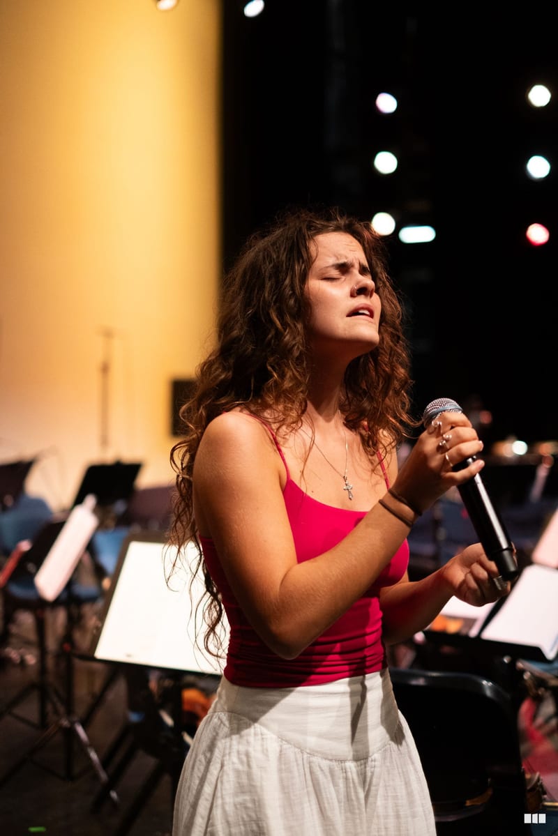 A young female soloist in a red top performs with a microphone in front of an orchestra during an evening concert.