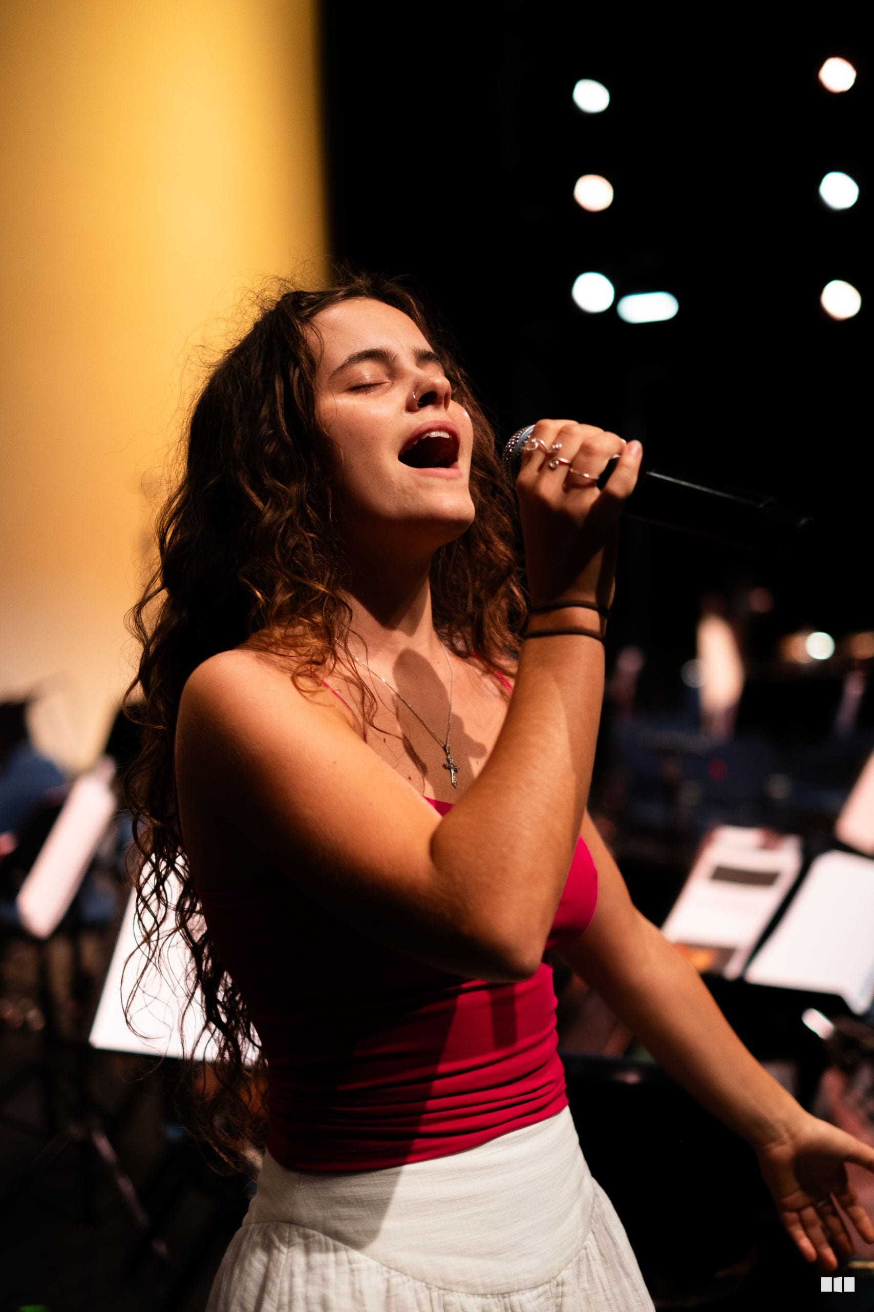 A female soloist performs passionately at the microphone under warm stage lighting with orchestra members visible in the background.