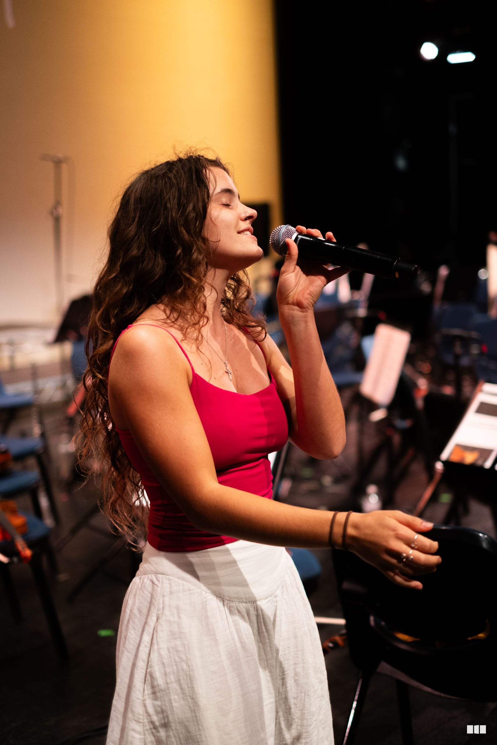 A female soloist in a red top and white skirt performs with a microphone against a warm golden stage backdrop.