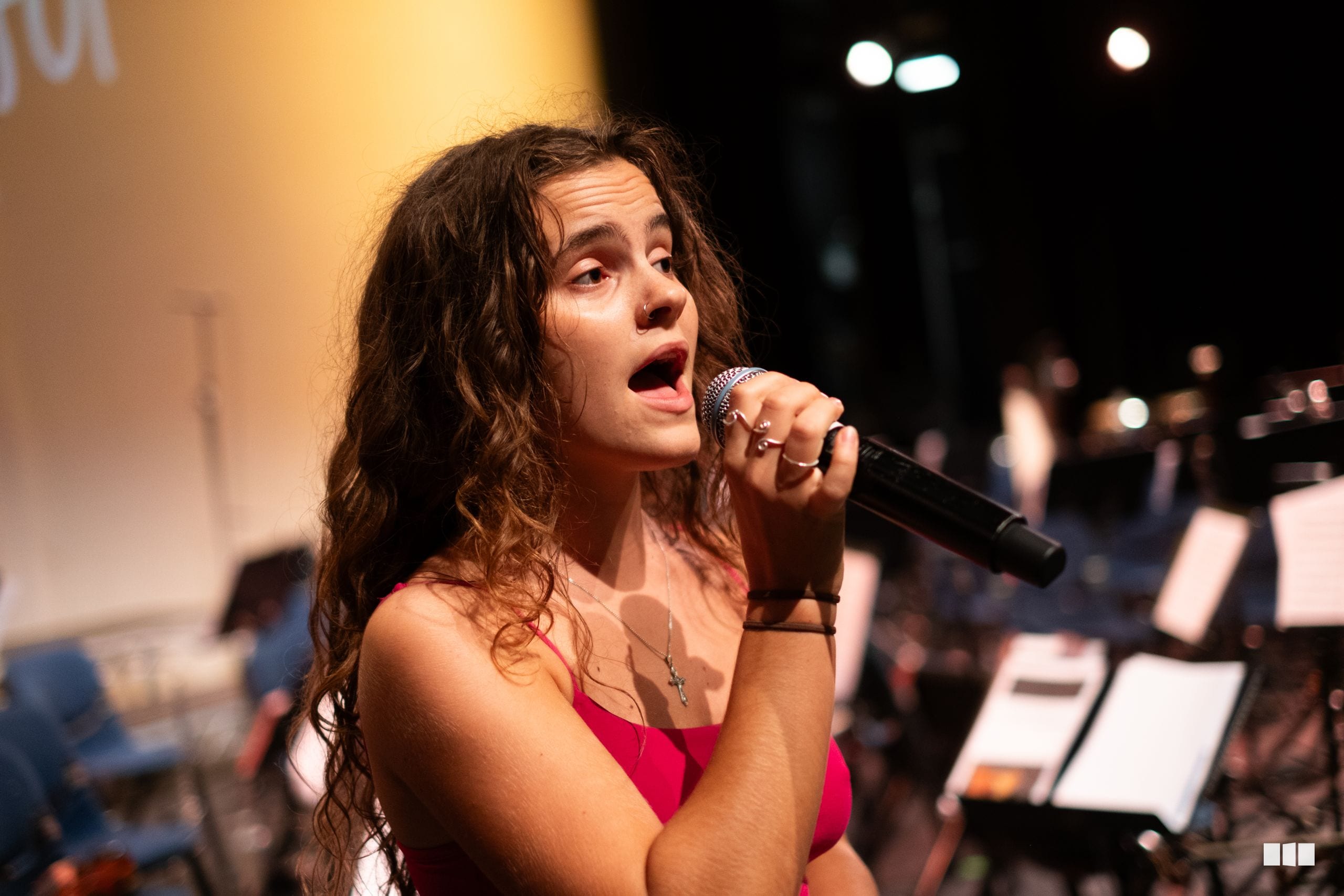 A young female soloist in a vibrant pink dress performs with a microphone, accompanied by musicians with sheet music visible in the background.