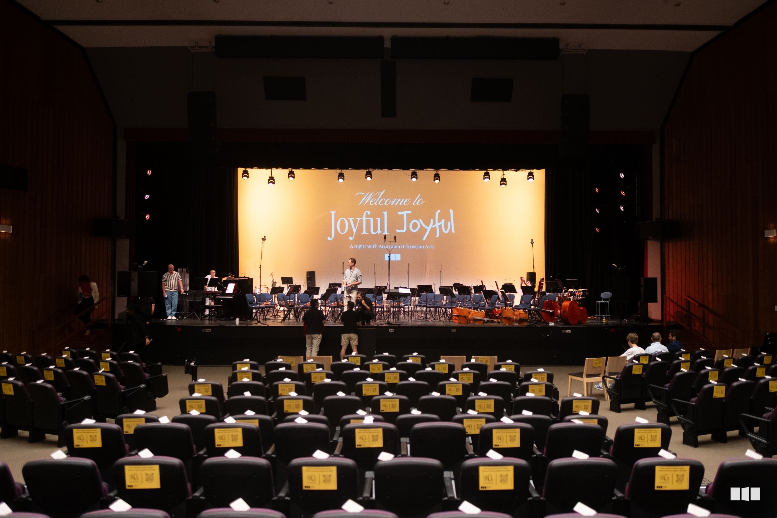 The Australian Christian Arts orchestra and ensemble perform during a concert titled 'Welcome to Joyful Joyful' in a large auditorium with reserved seating.