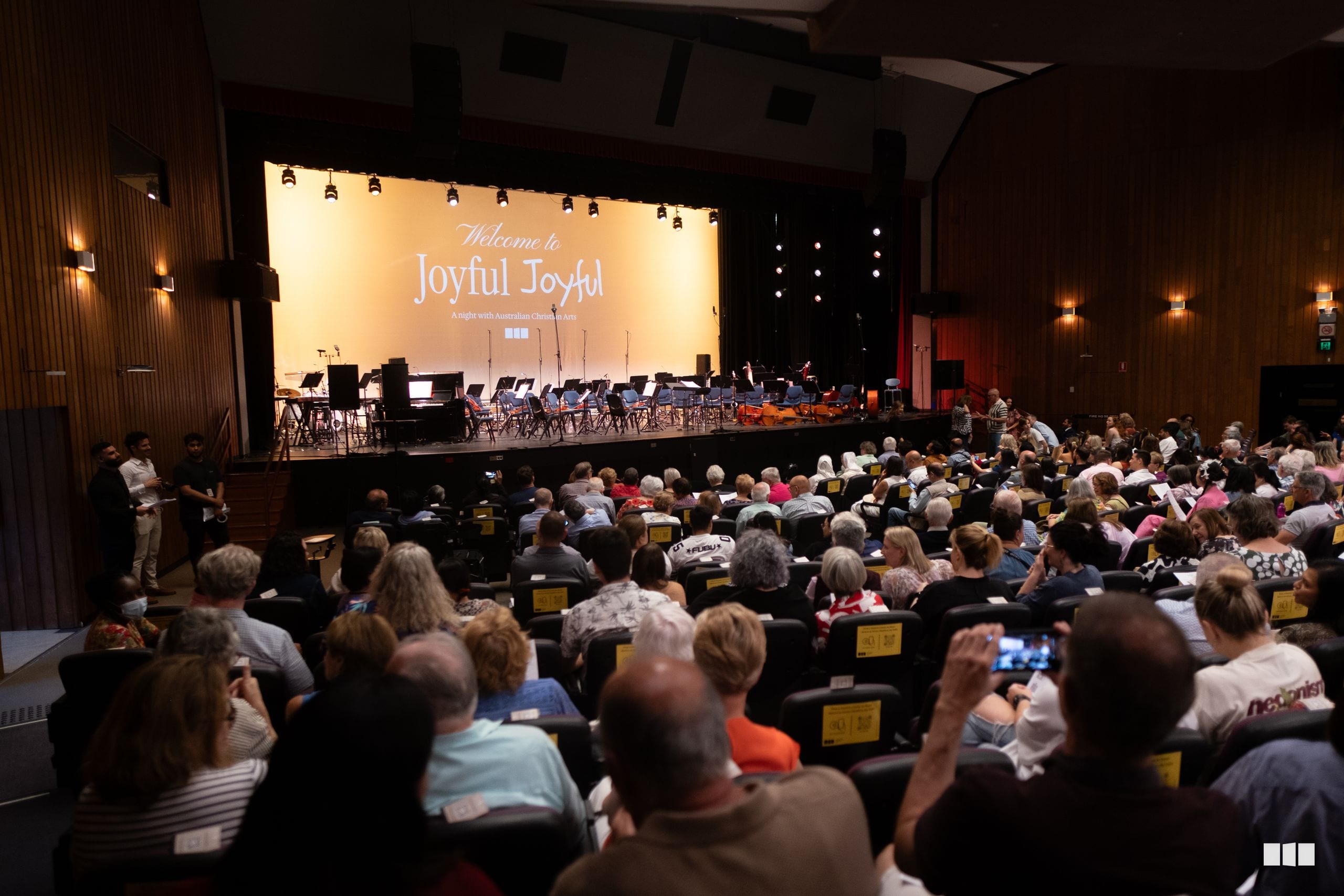 A full auditorium of audience members watches the Australian Christian Arts orchestra and choir perform during the 'Joyful Joyful' concert event on stage.