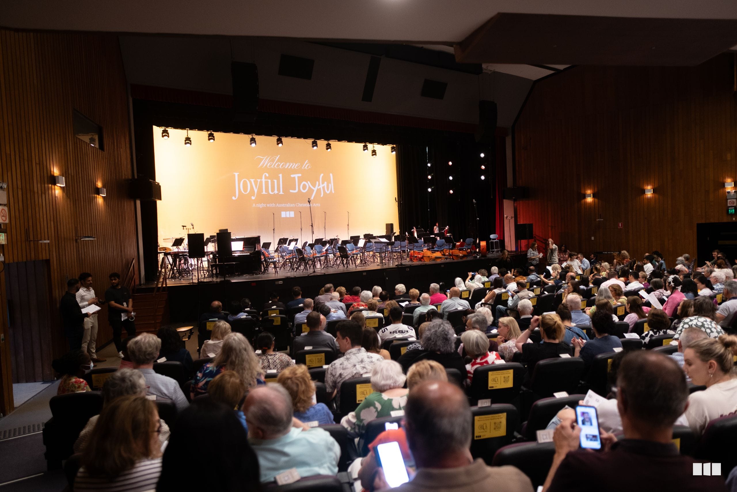 A large audience fills the auditorium as the orchestra and choir perform on stage during the 'Joyful Joyful' concert.