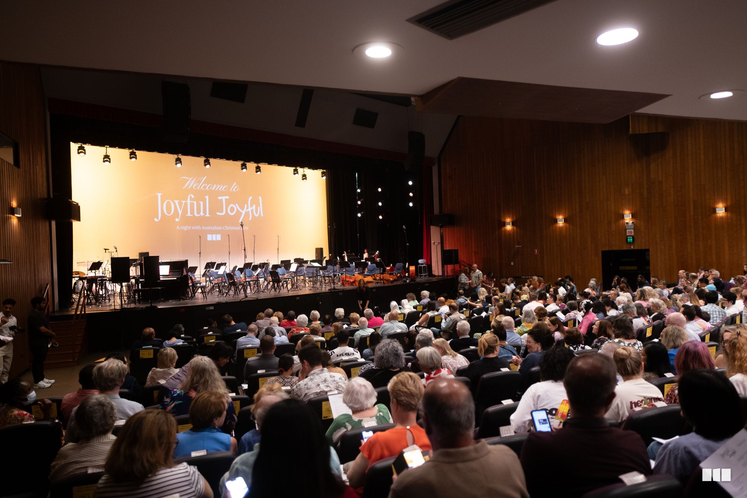 A large audience watches an orchestra and choir perform on stage at an Australian Christian Arts concert titled 'Welcome to Joyful Joyful.'