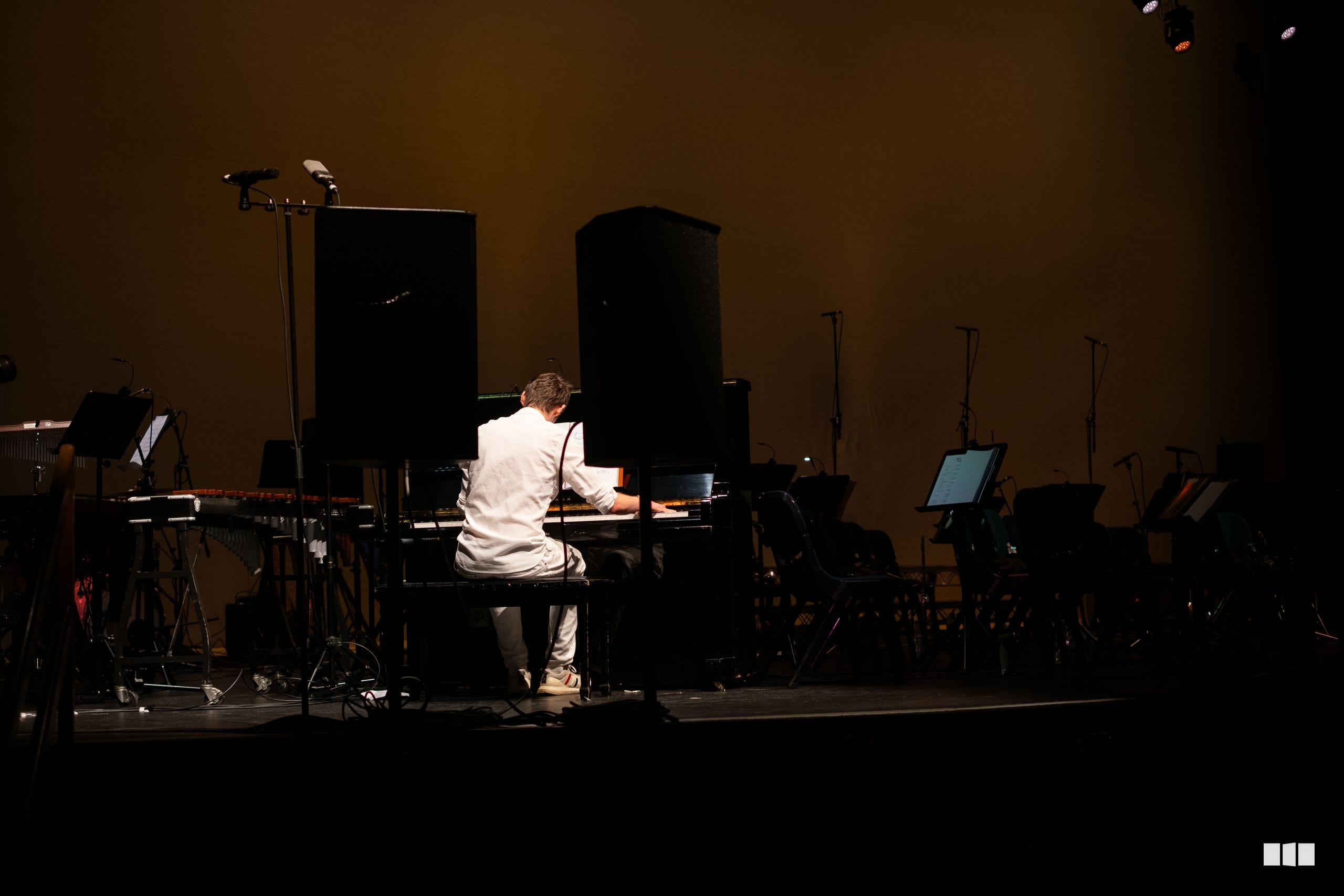 A pianist in a white shirt performs at a grand piano on stage with orchestral musicians and equipment visible in the background.