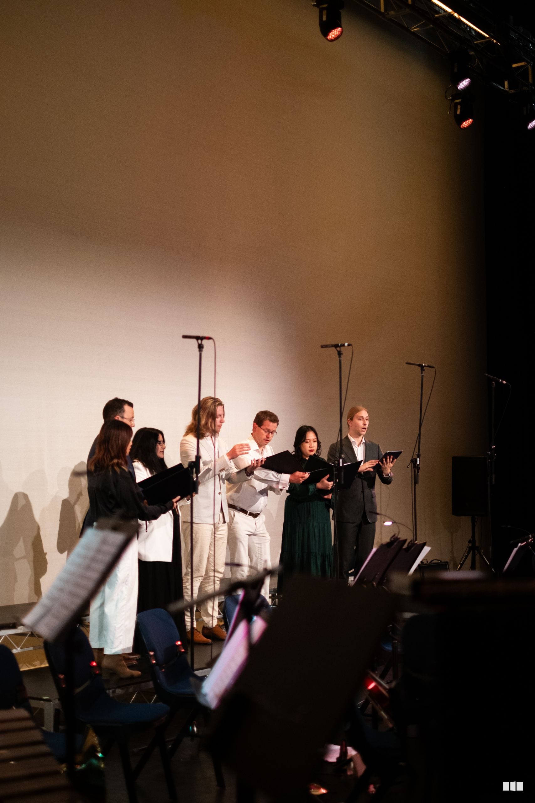 A choir ensemble performs on stage with sheet music, illuminated by stage lighting during a choral performance.