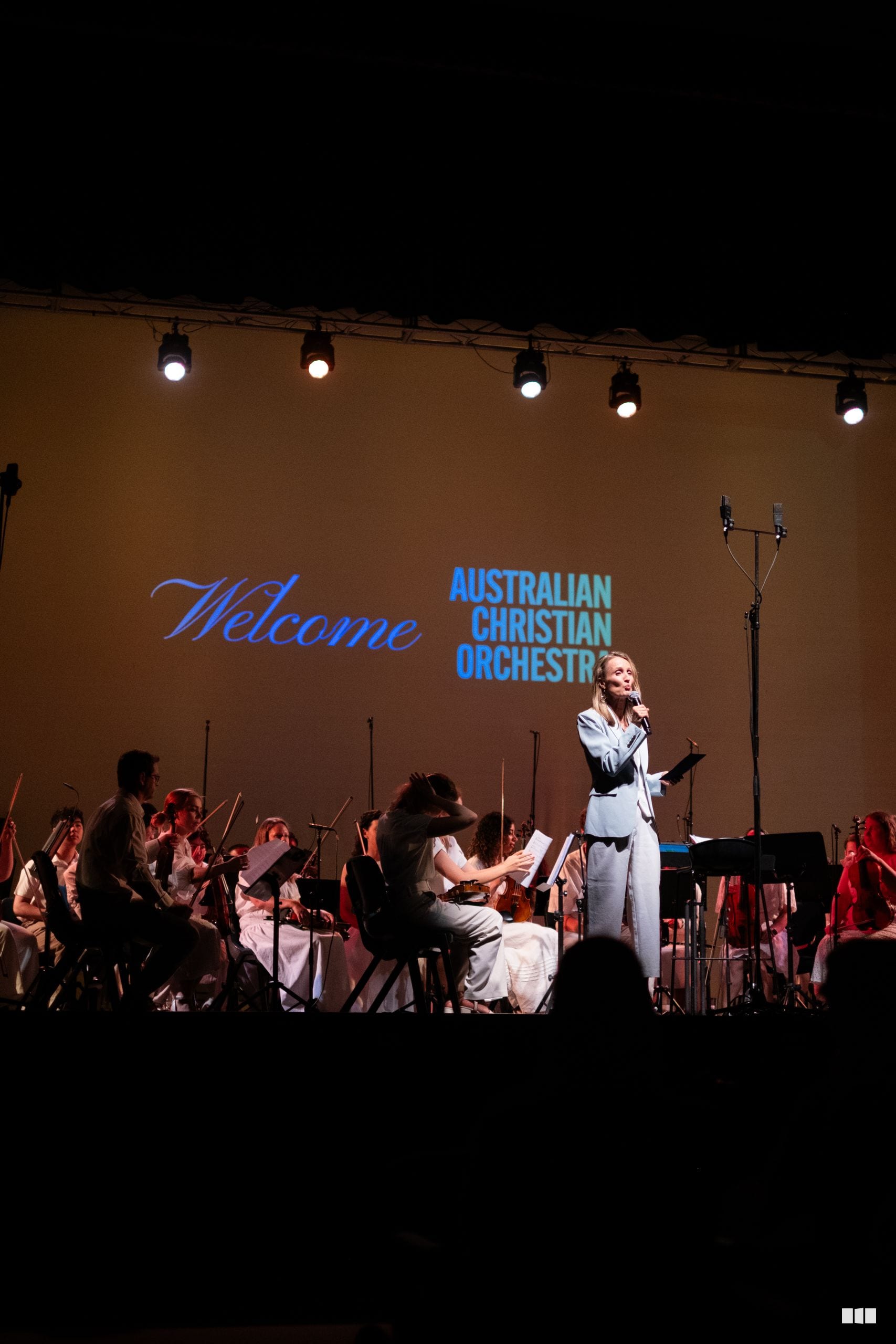 A female soloist in a light blue jacket addresses the Australian Christian Orchestra during a welcome performance on stage.