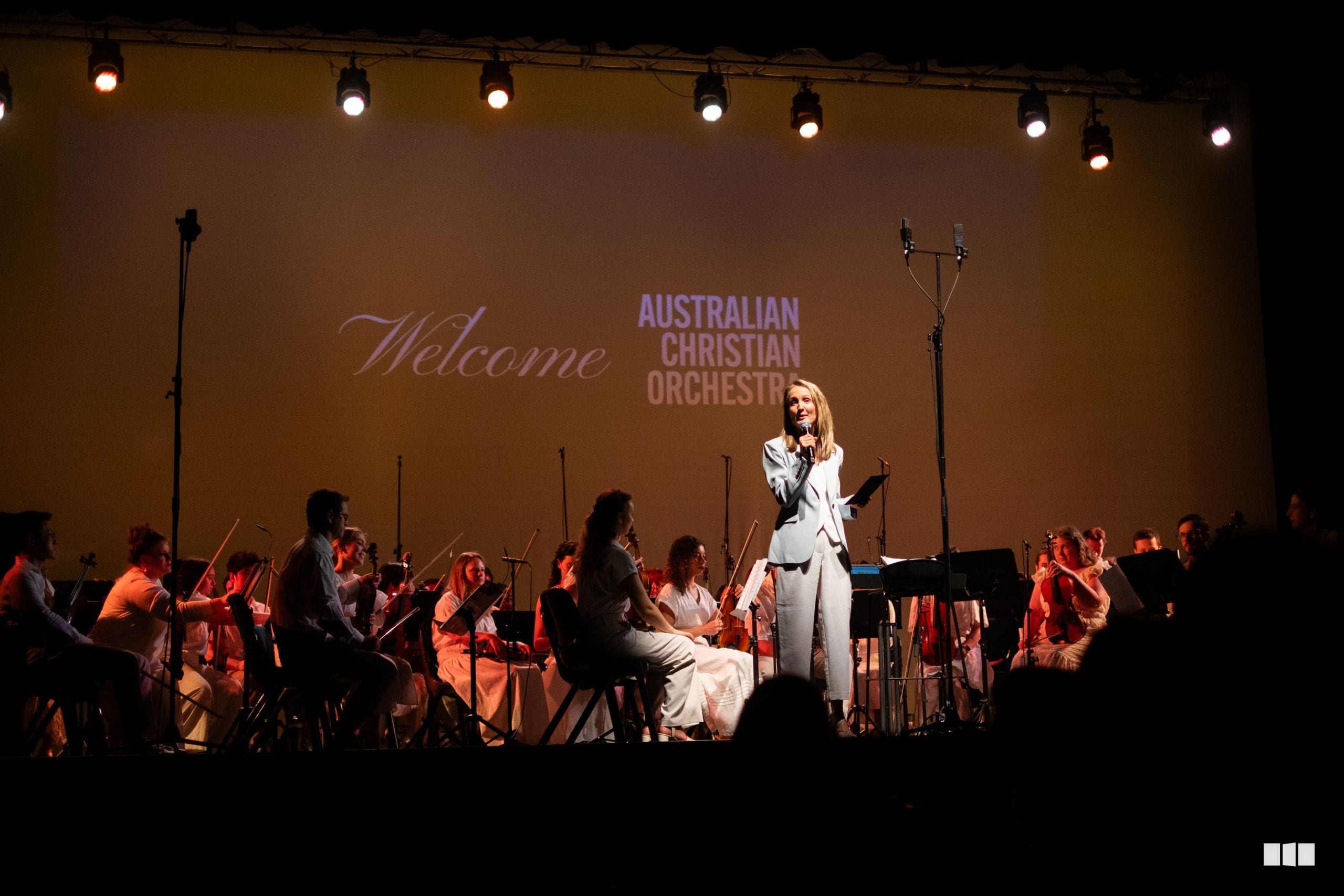 A female soloist in white performs at the microphone with the Australian Christian Orchestra accompanying her on stage.