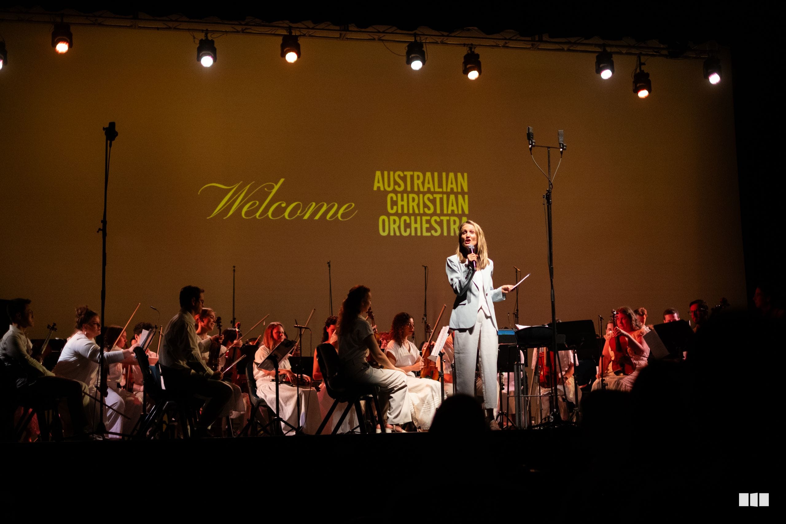 A soloist in white performs at the center stage with the Australian Christian Orchestra seated around her during a concert event.
