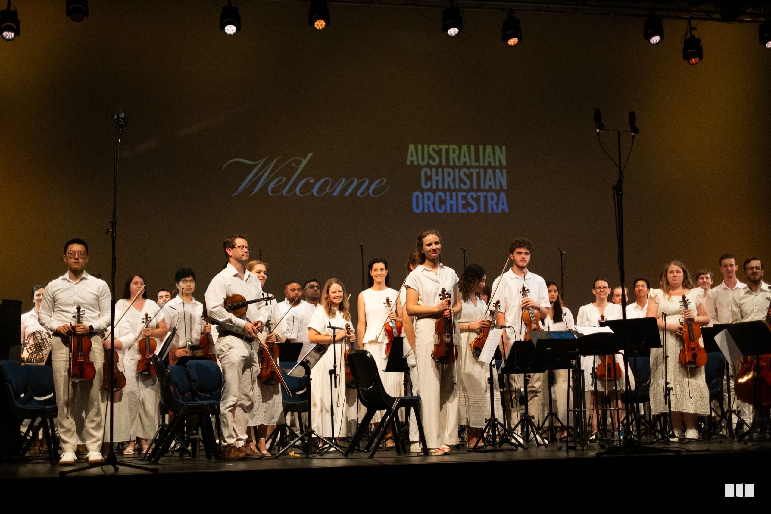 The Australian Christian Orchestra ensemble takes a bow on stage following their performance, with musicians dressed in white attire.