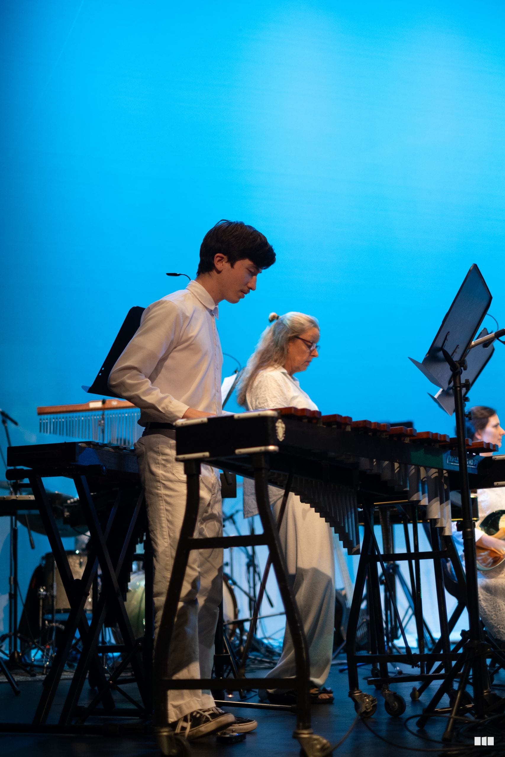 Young percussionist performs at a vibraphone alongside an older musician on stage against a bright blue backdrop.
