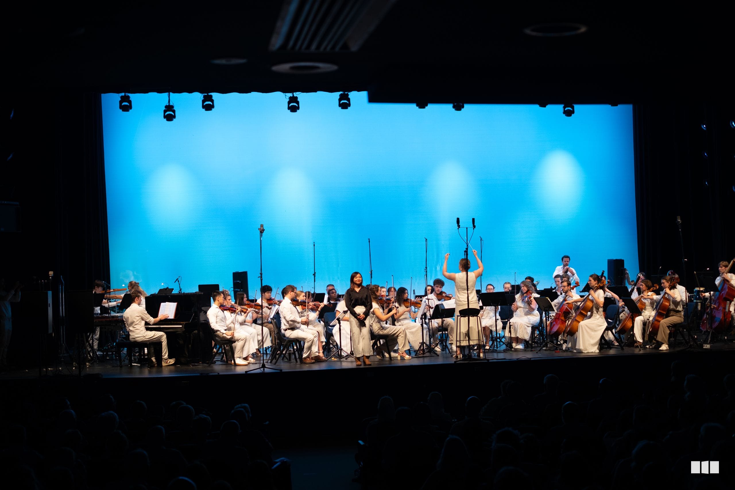 A conductor leads a full orchestra in white attire against a vibrant blue illuminated backdrop during a concert performance.