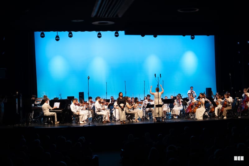 A conductor leads a full orchestra in white attire against a vibrant blue illuminated backdrop during a concert performance.