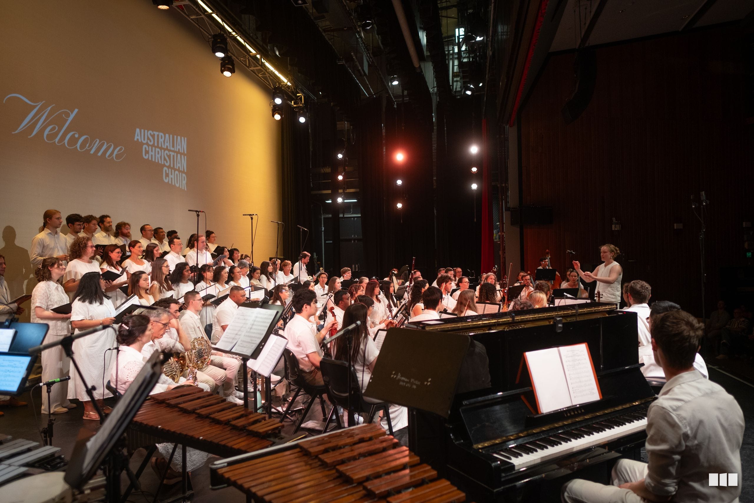 The Australian Christian Choir performs with orchestra members and piano accompaniment on stage during a concert.