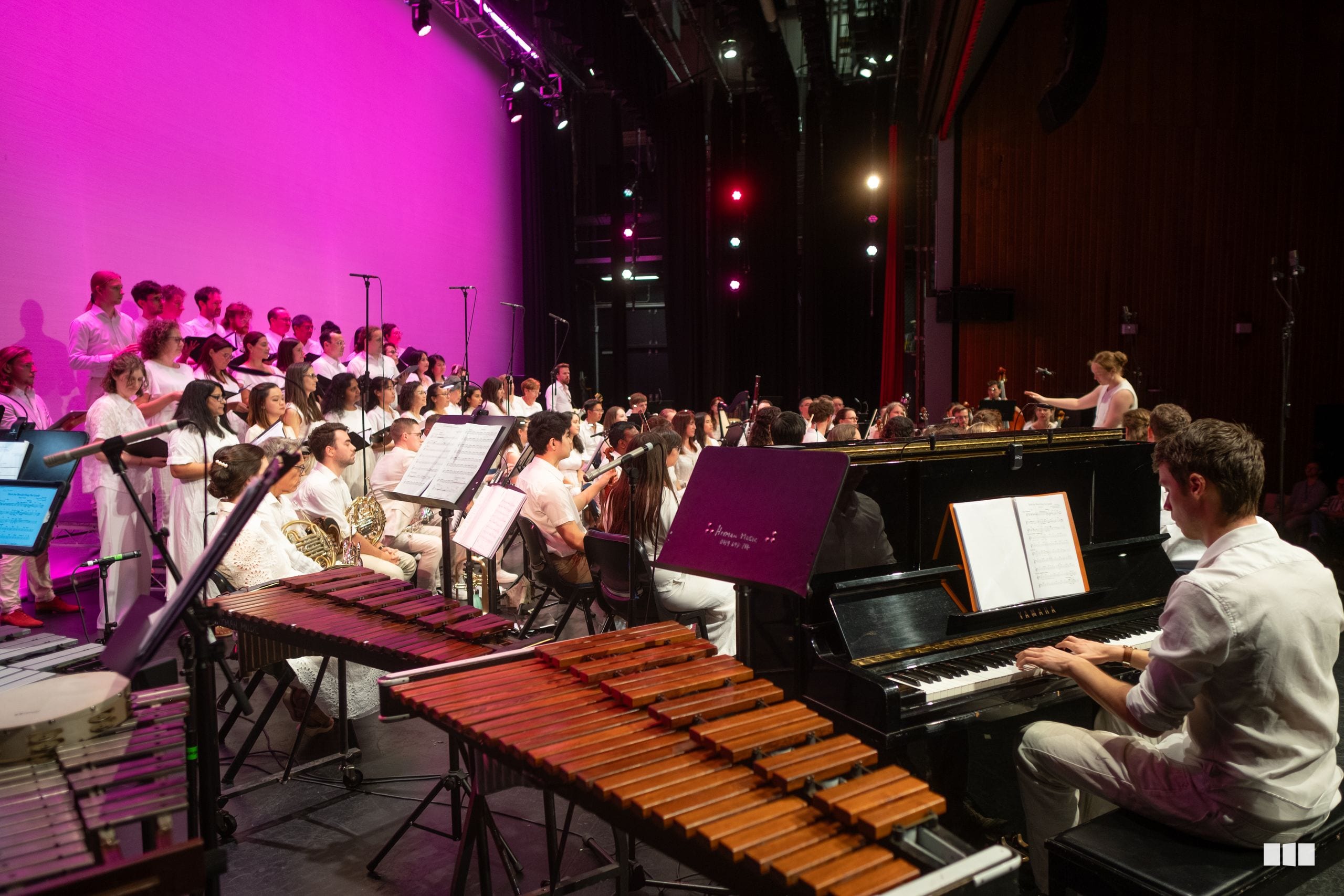 A conductor leads a combined orchestra and choir performance featuring vibraphone and marimba soloists on stage with pink lighting and multiple instrumental sections.