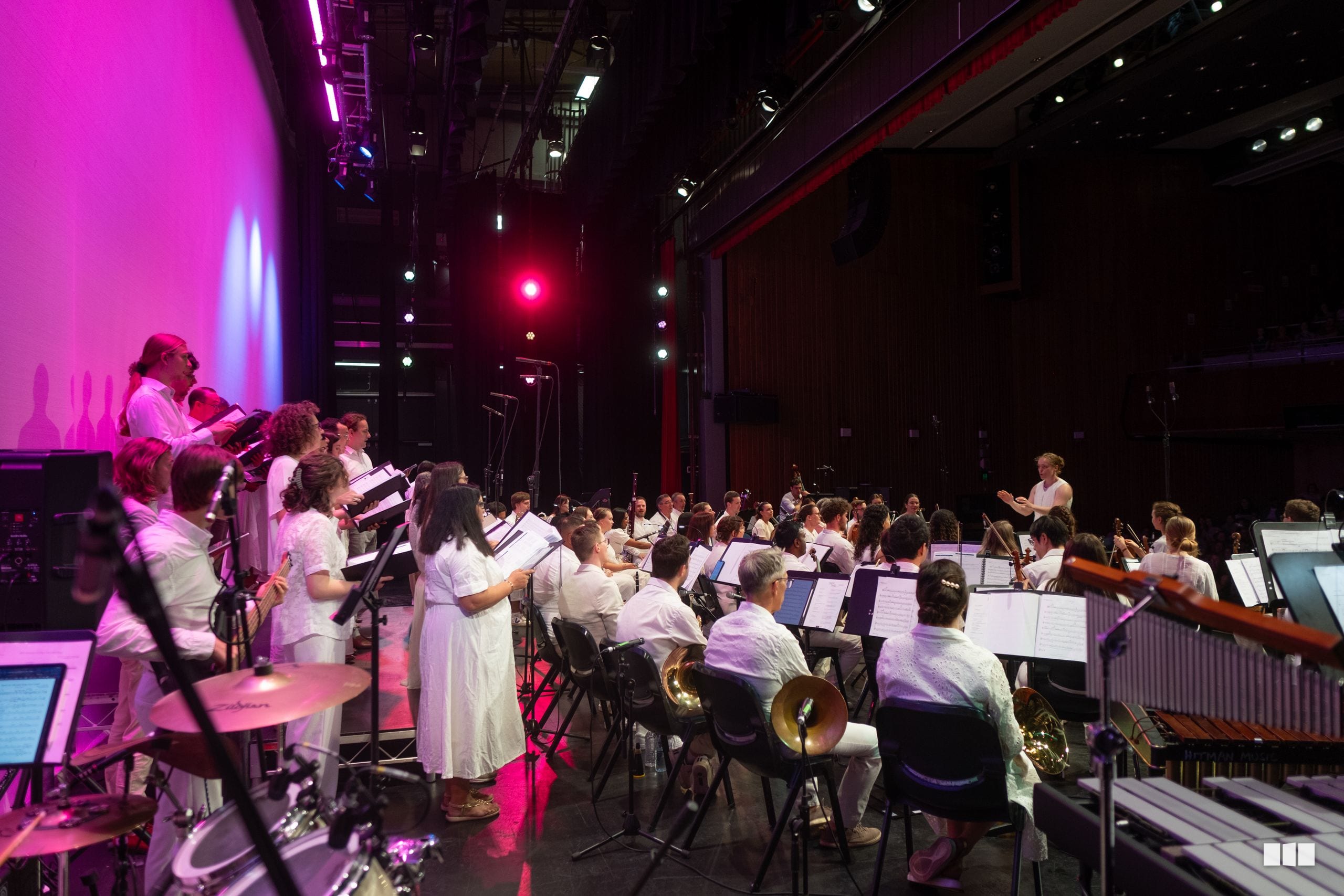 A conductor leads a full orchestra and choir in a concert performance under vibrant pink and red stage lighting.