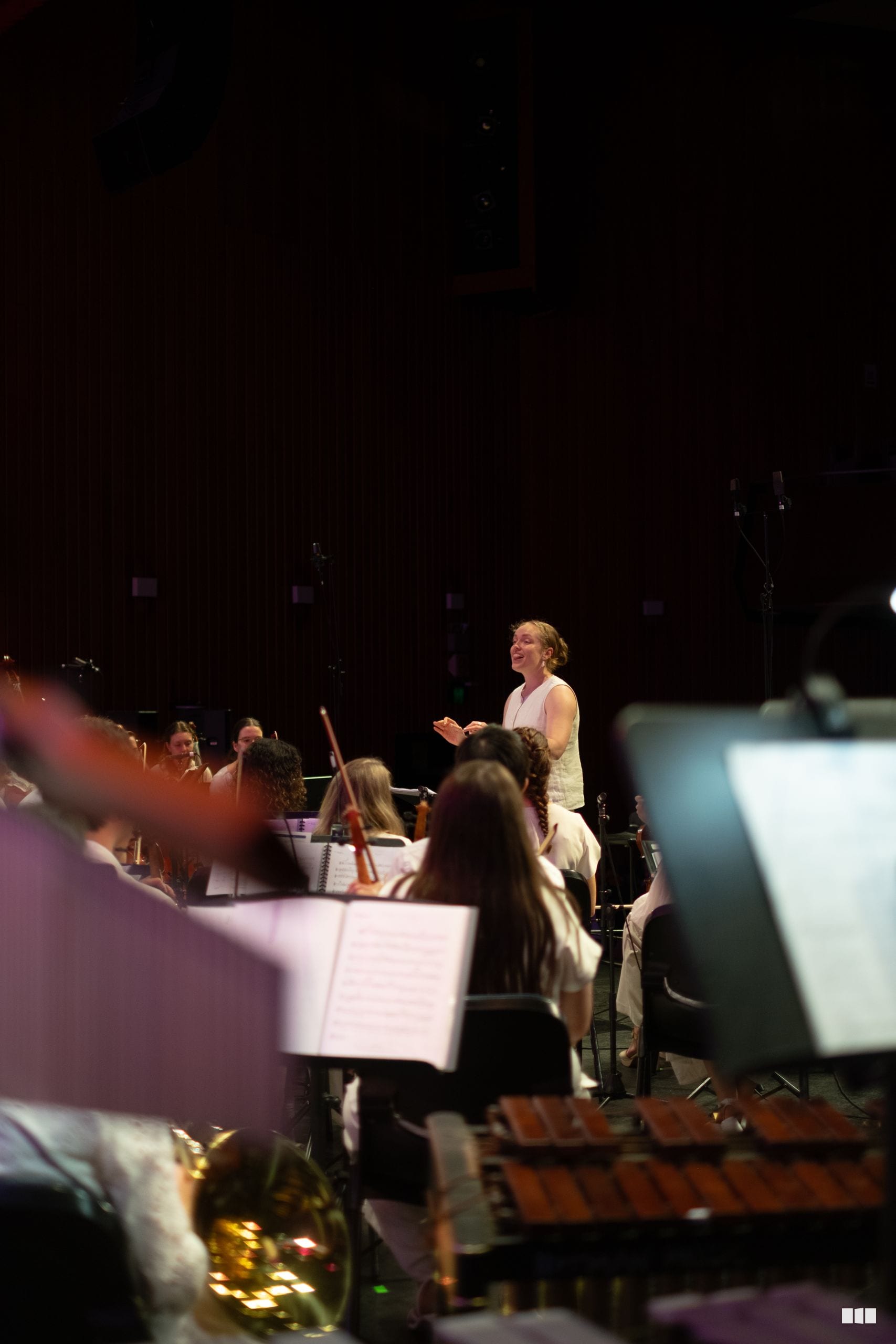 A conductor leads an orchestra during a concert performance, with musicians holding sheet music in the foreground.