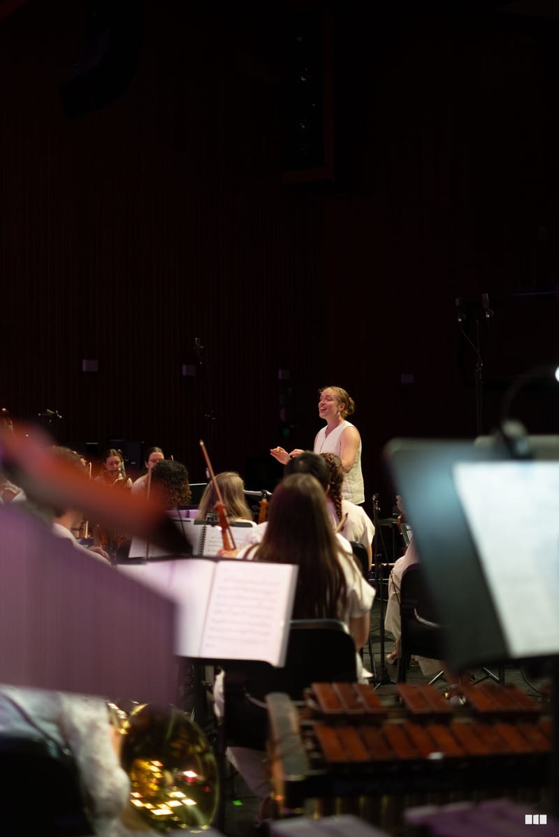 A conductor leads an orchestra during a concert performance, with musicians holding sheet music in the foreground.
