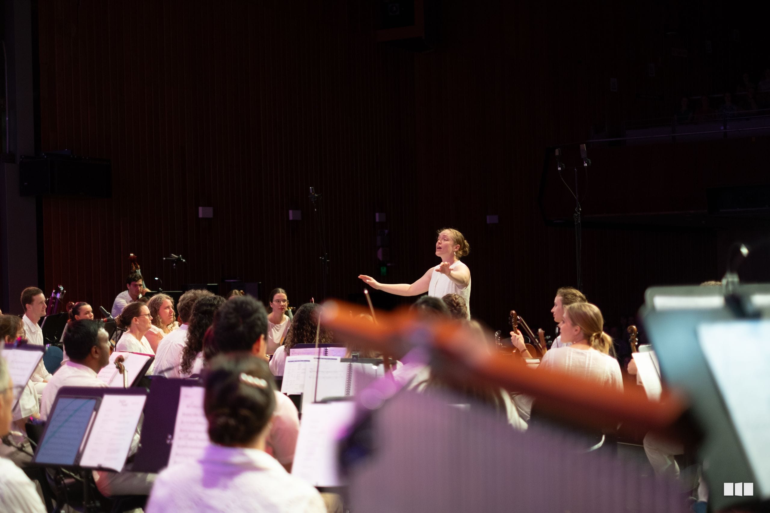 A conductor leads an orchestra during a rehearsal or performance, with musicians seated with their sheet music under stage lighting.