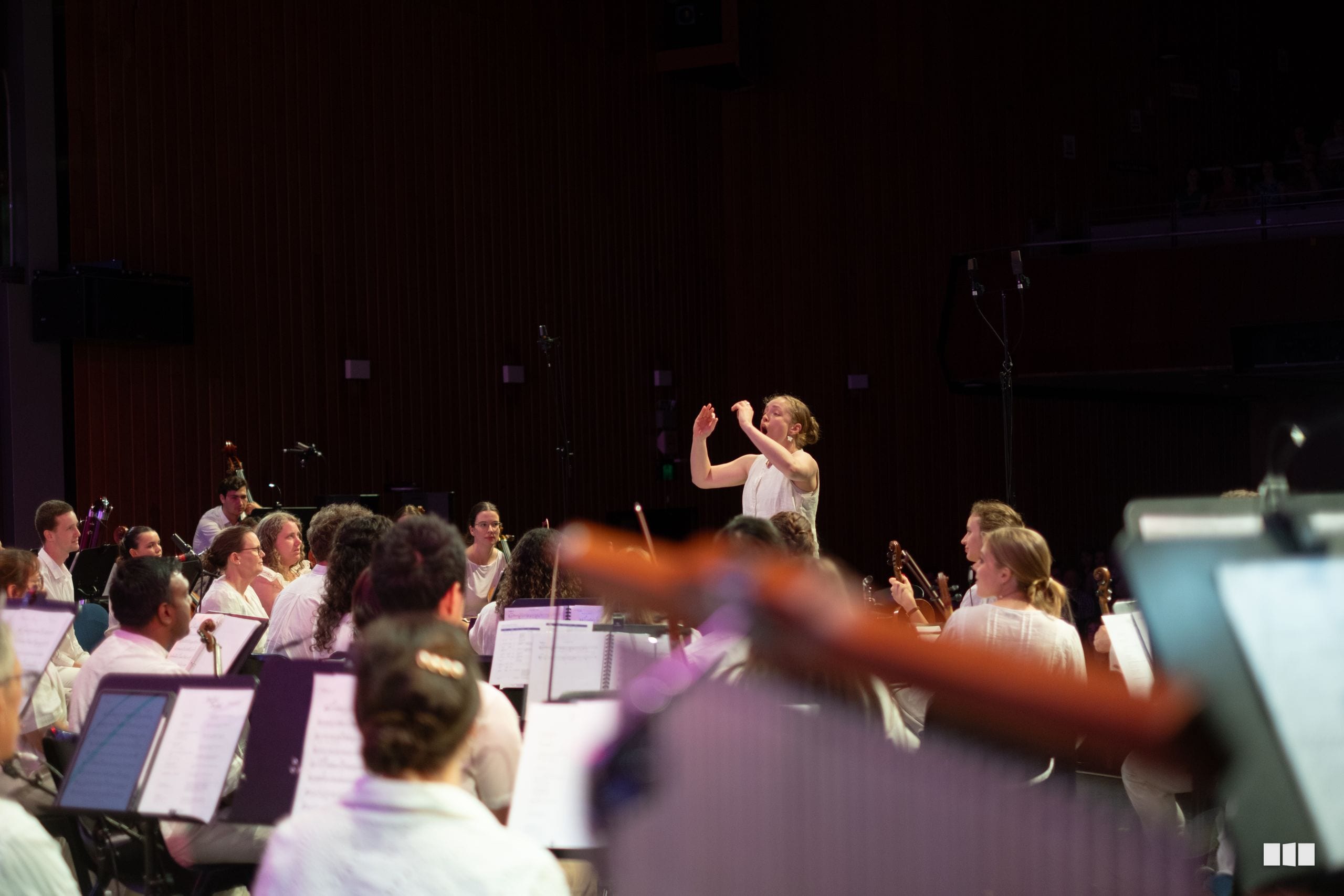 A conductor leads an orchestra during rehearsal in a concert hall with sheet music and musicians visible in the foreground.