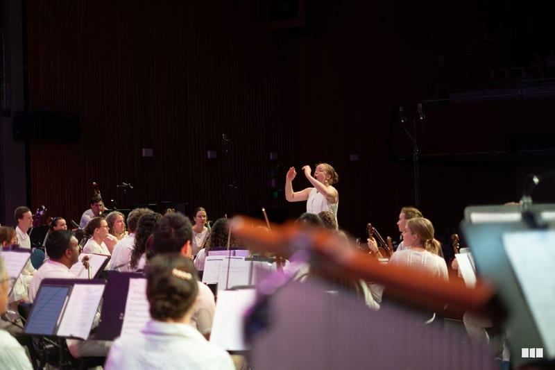 A conductor leads an orchestra during rehearsal in a concert hall with sheet music and musicians visible in the foreground.
