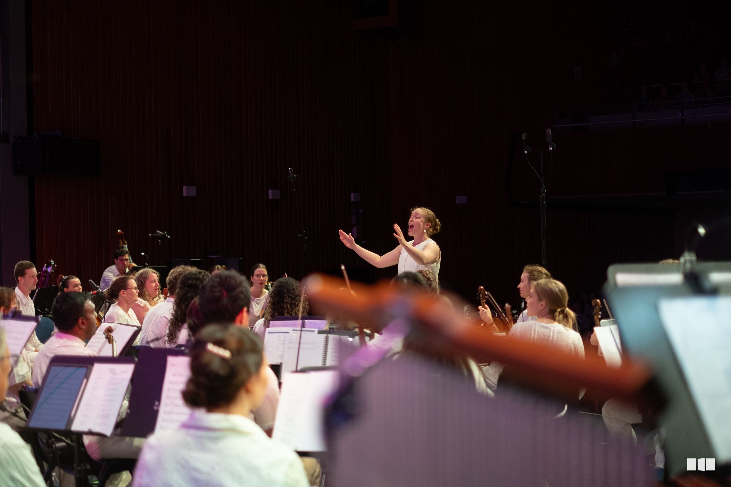 A conductor leads an orchestra during a rehearsal or performance, with musicians holding sheet music and instruments in the foreground.