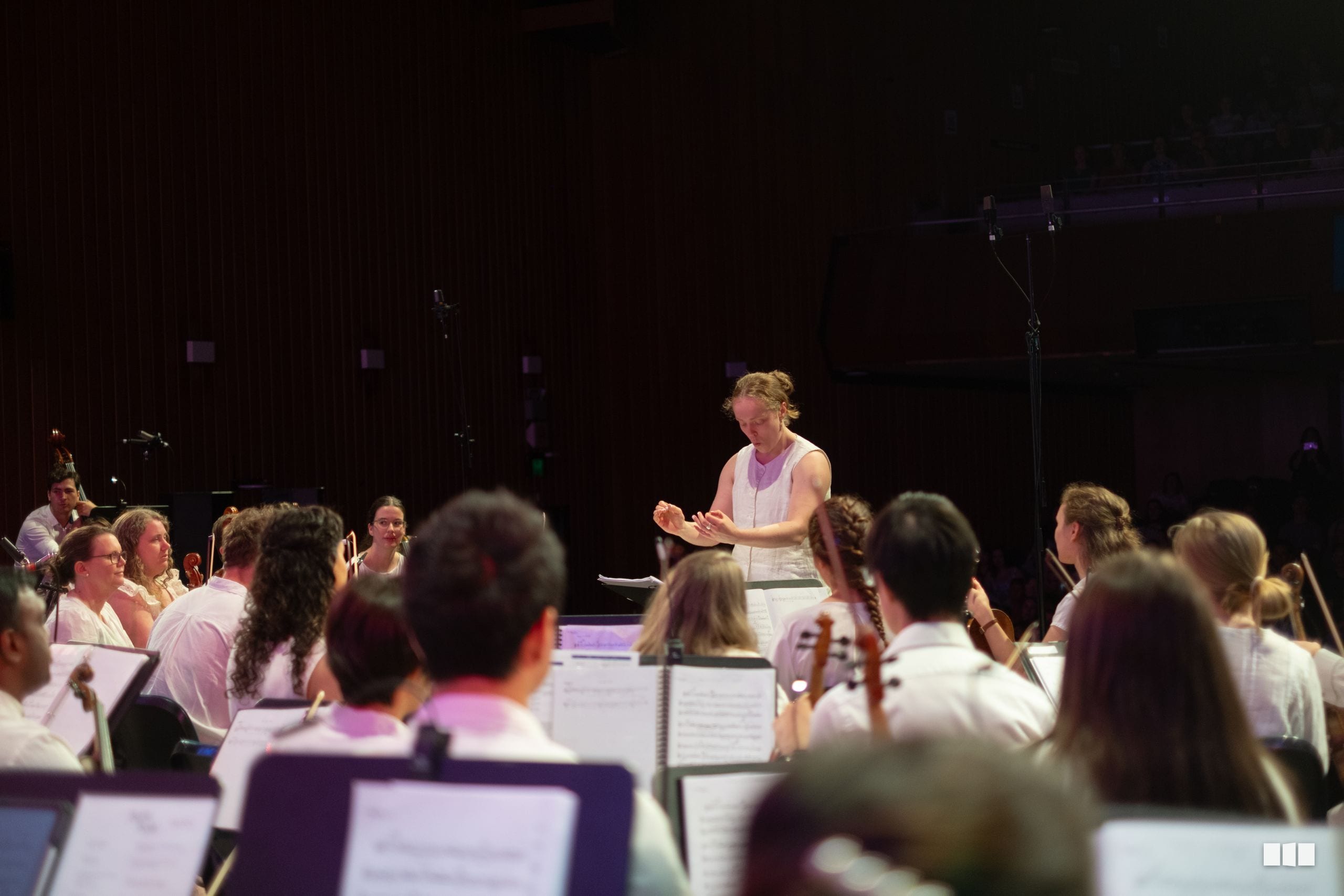 A conductor leads an orchestra during a rehearsal or performance, with musicians holding instruments and sheet music visible in the foreground.
