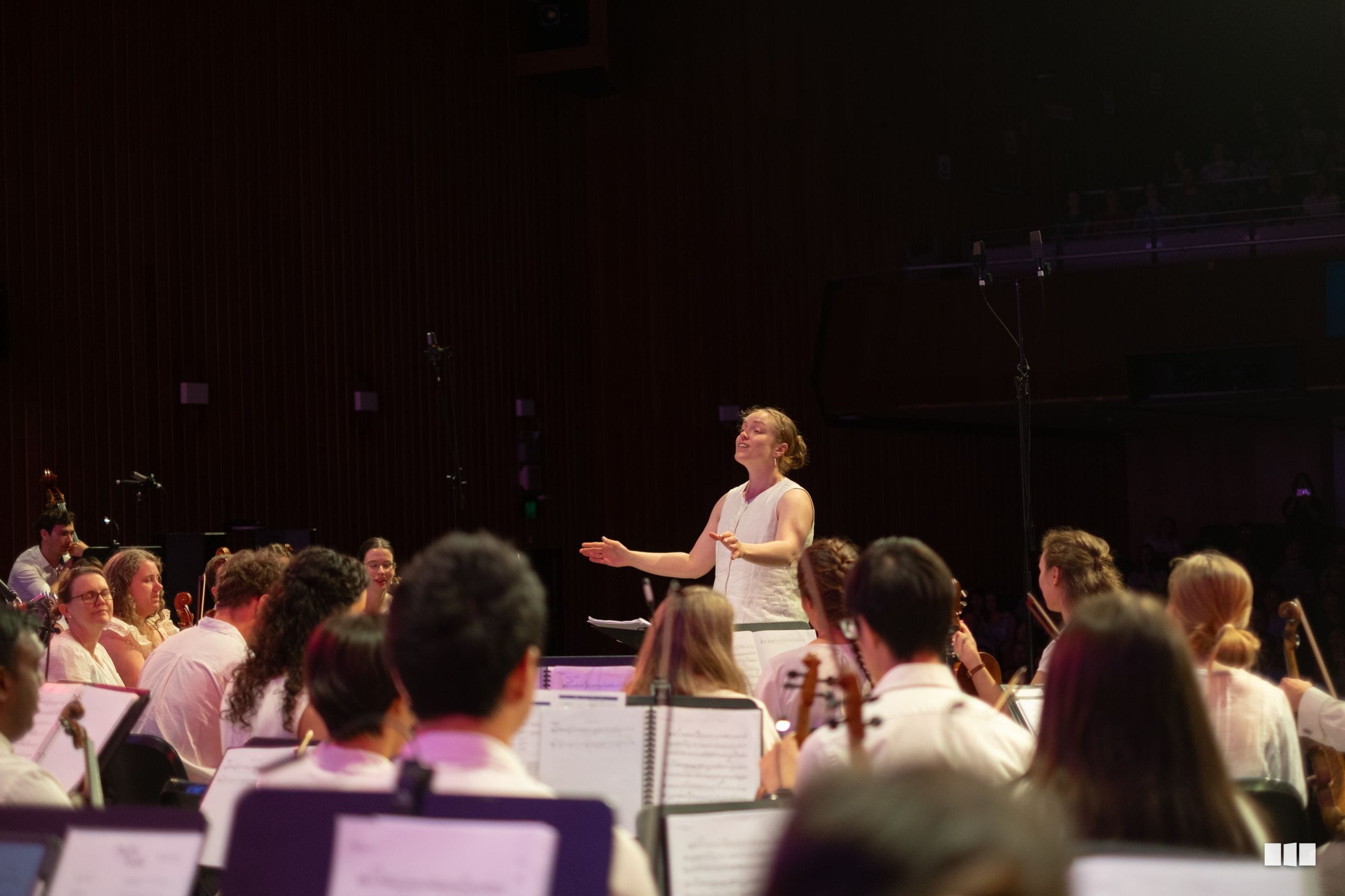 A conductor leads an orchestra during a rehearsal or performance, with musicians seated with sheet music in the foreground.