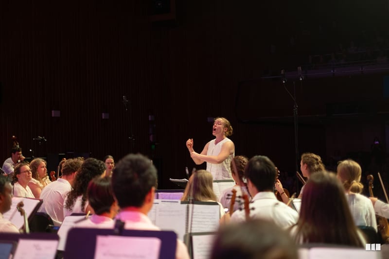 A conductor leads an orchestra and choir during a rehearsal or performance in a concert hall.