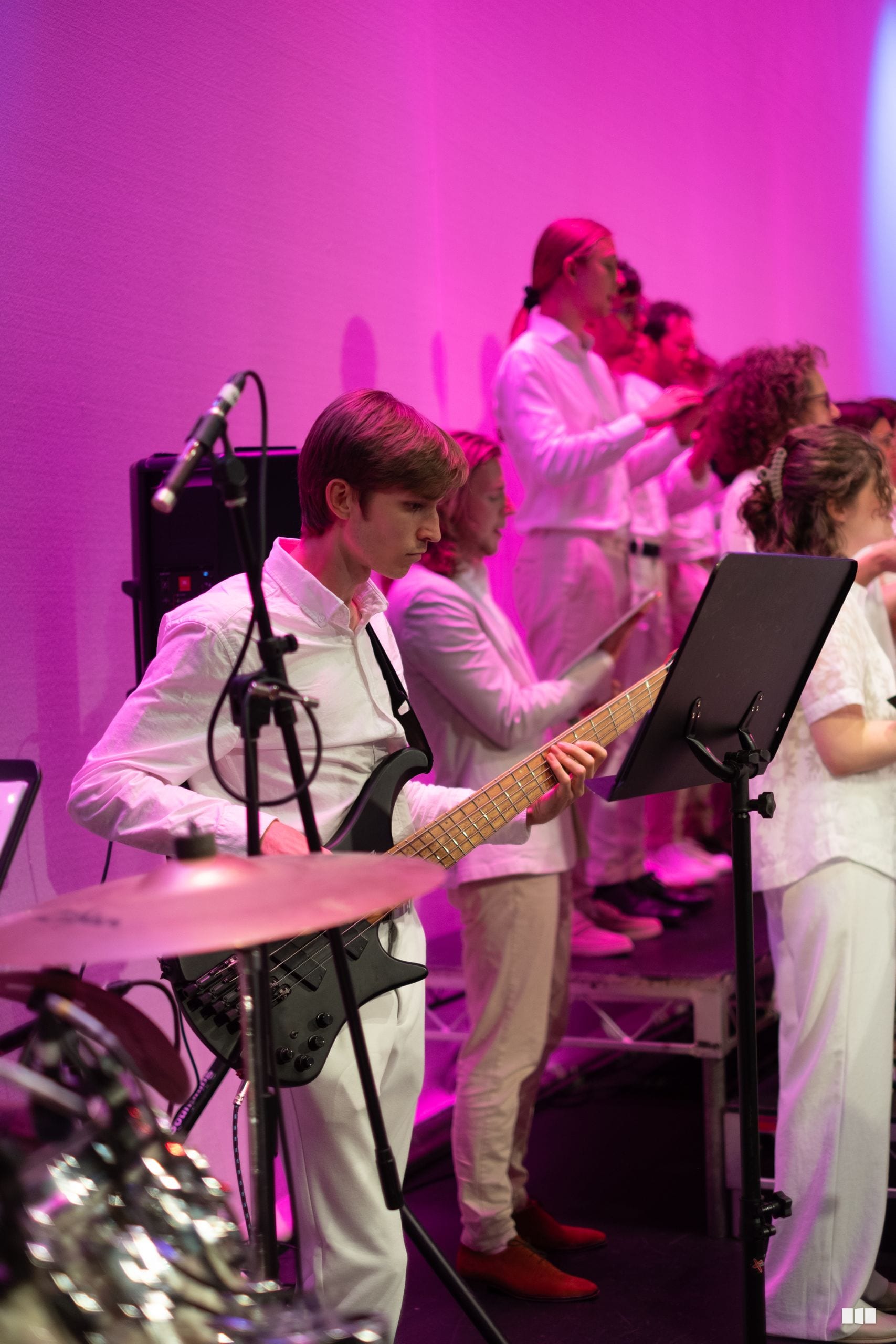 Young bass guitarist performs with sheet music under vibrant pink stage lighting during an ensemble performance.
