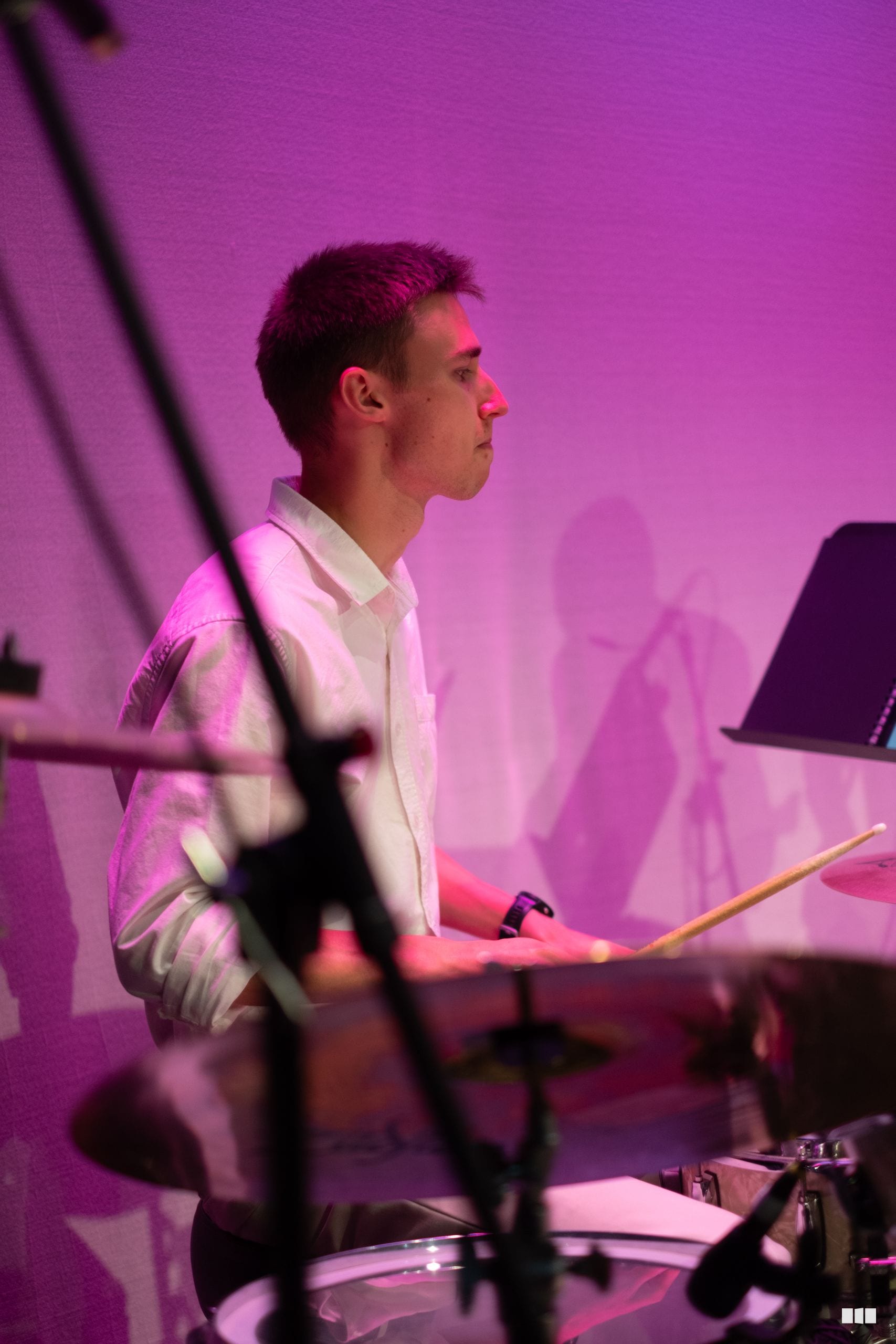 A young drummer performs at the kit during an orchestral concert under vibrant purple stage lighting.