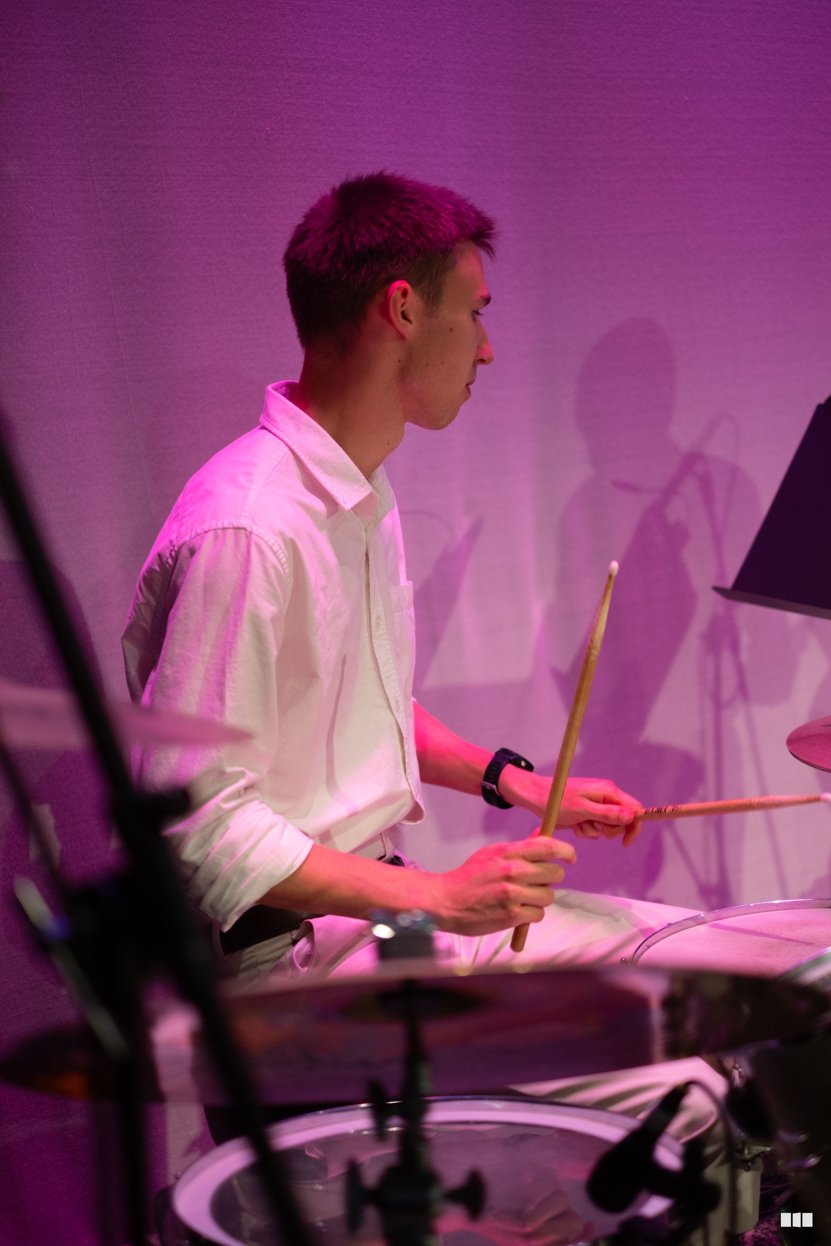 A young drummer performs at the drums during an orchestral concert, illuminated by pink stage lighting.