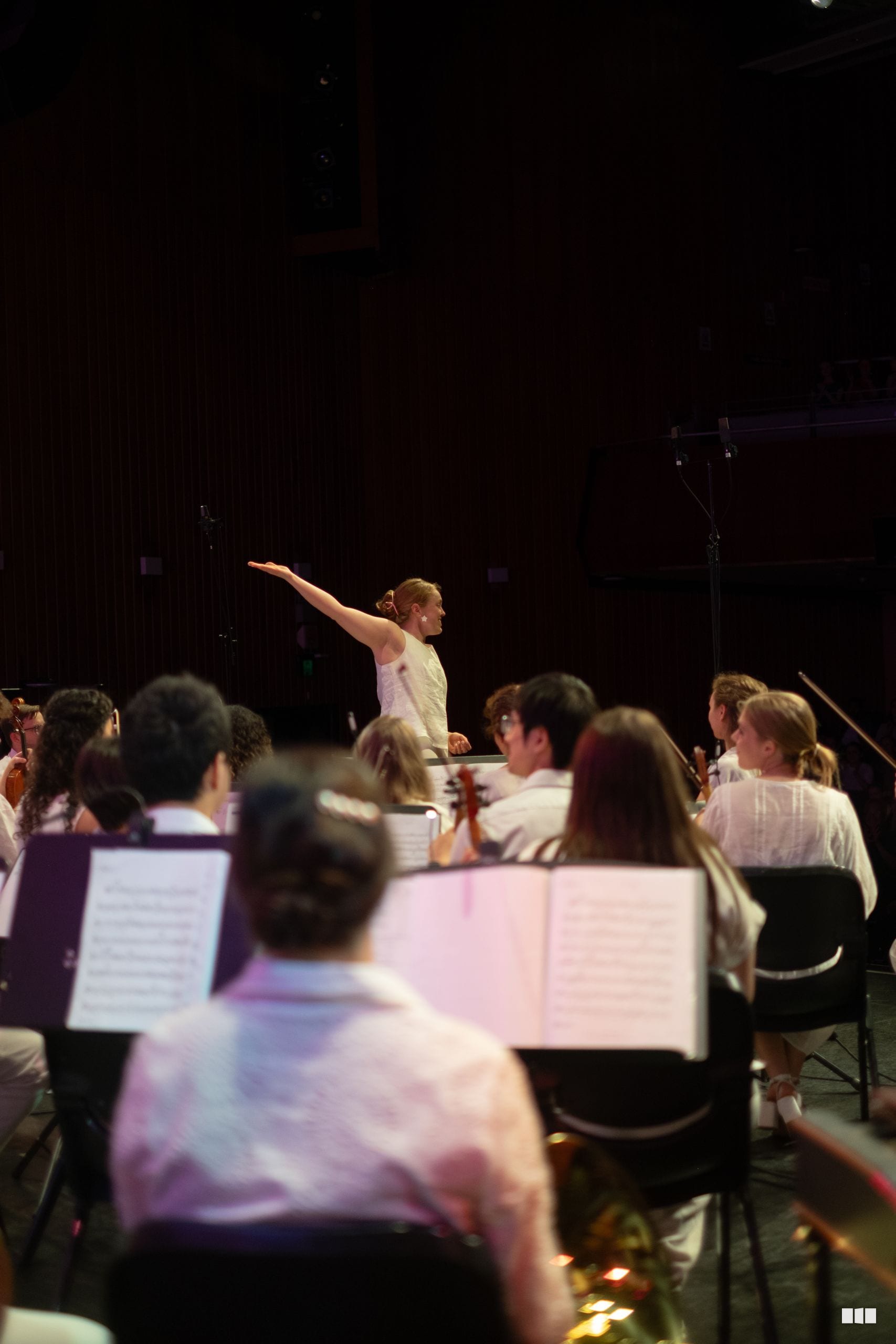 A conductor leads an orchestra during a rehearsal or performance, with musicians seated with sheet music in the foreground.