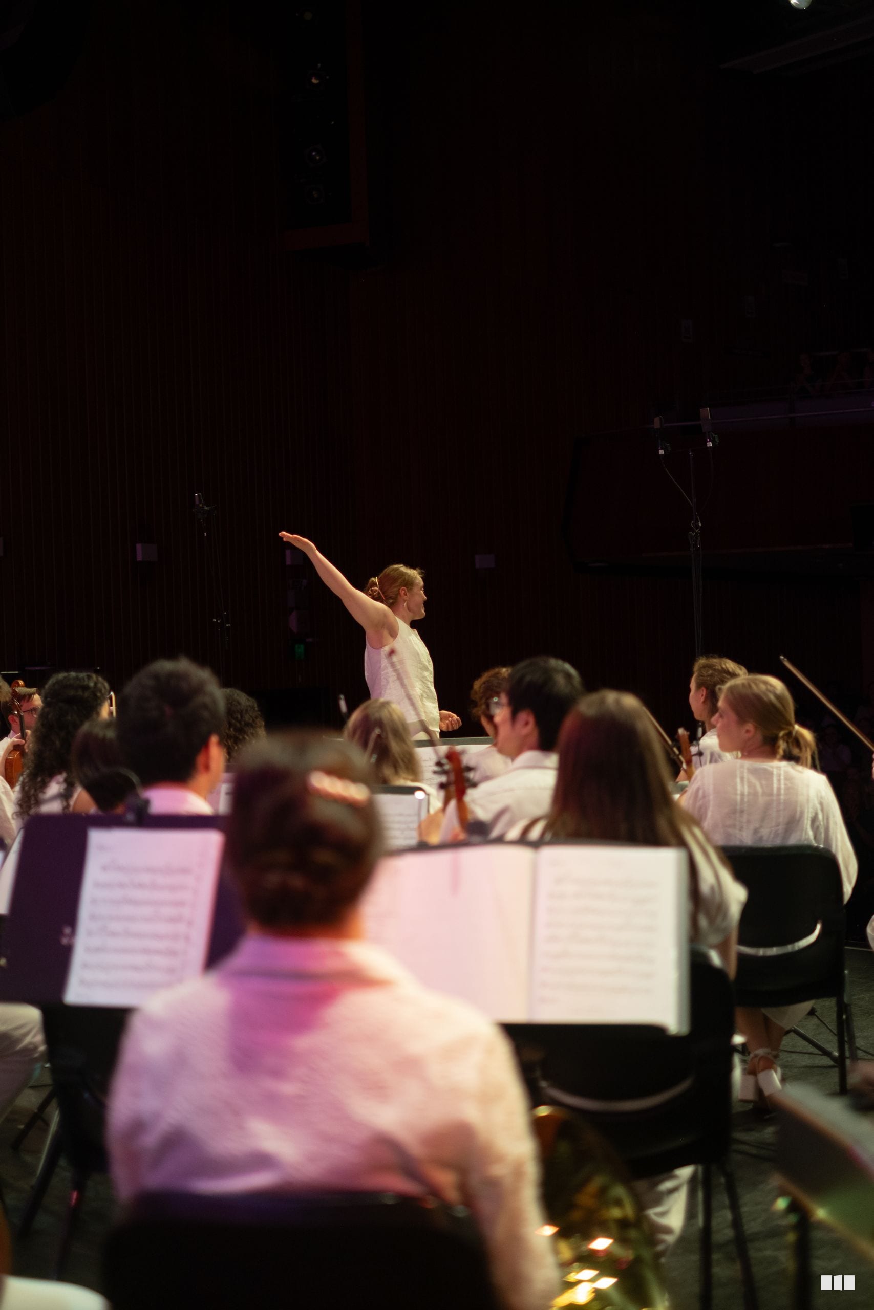 A conductor leads an orchestra during a rehearsal or performance, with musicians reading sheet music in the foreground.