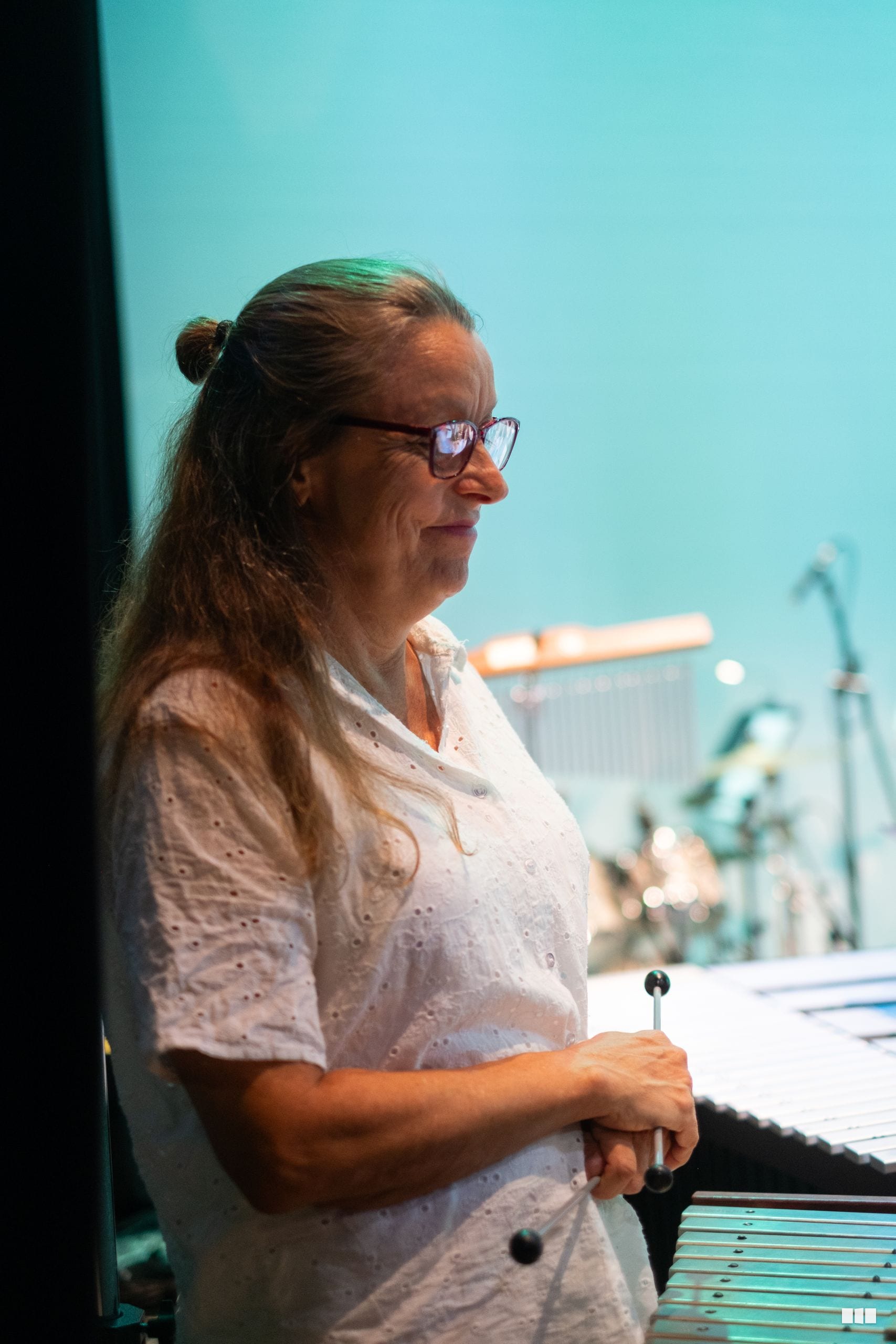 A woman wearing glasses and a white shirt stands at a piano keyboard during what appears to be a rehearsal or performance setting.