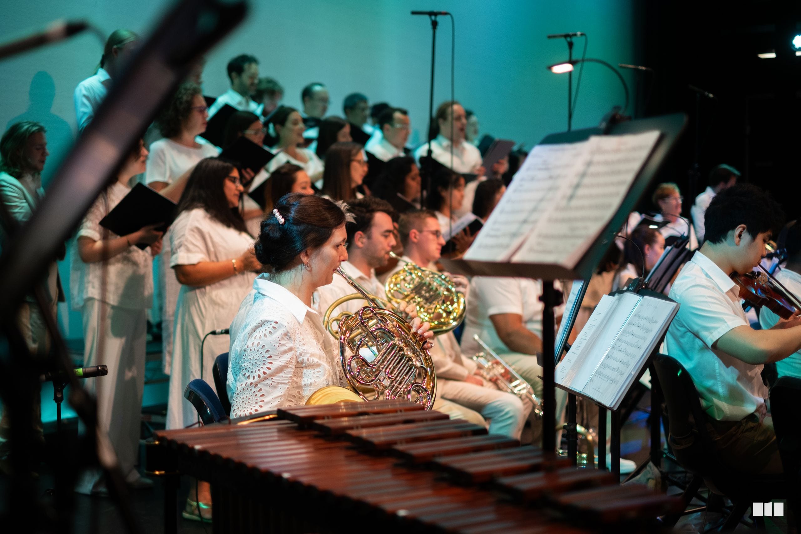 Orchestra and choir members perform together on stage with instrumental soloists and singers under turquoise stage lighting.