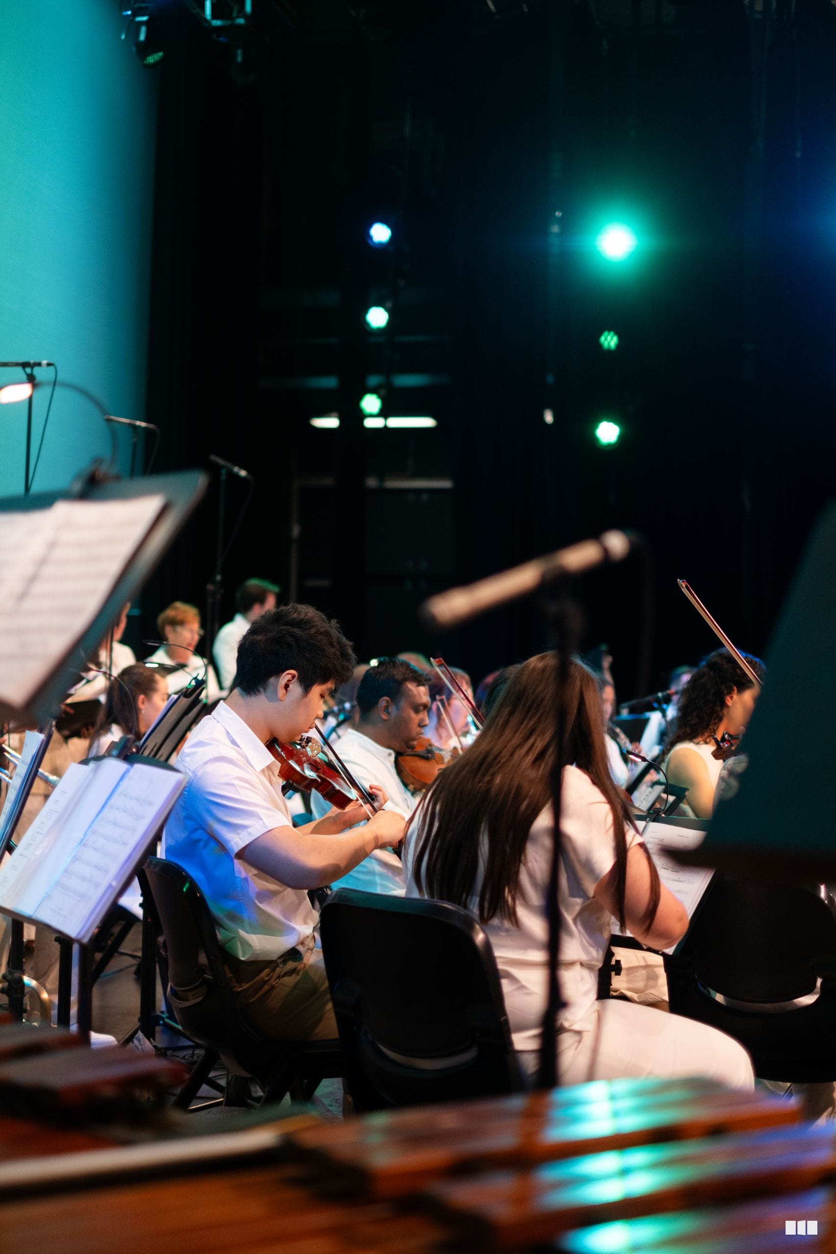 Young orchestra members perform with sheet music under stage lighting during a rehearsal or concert.