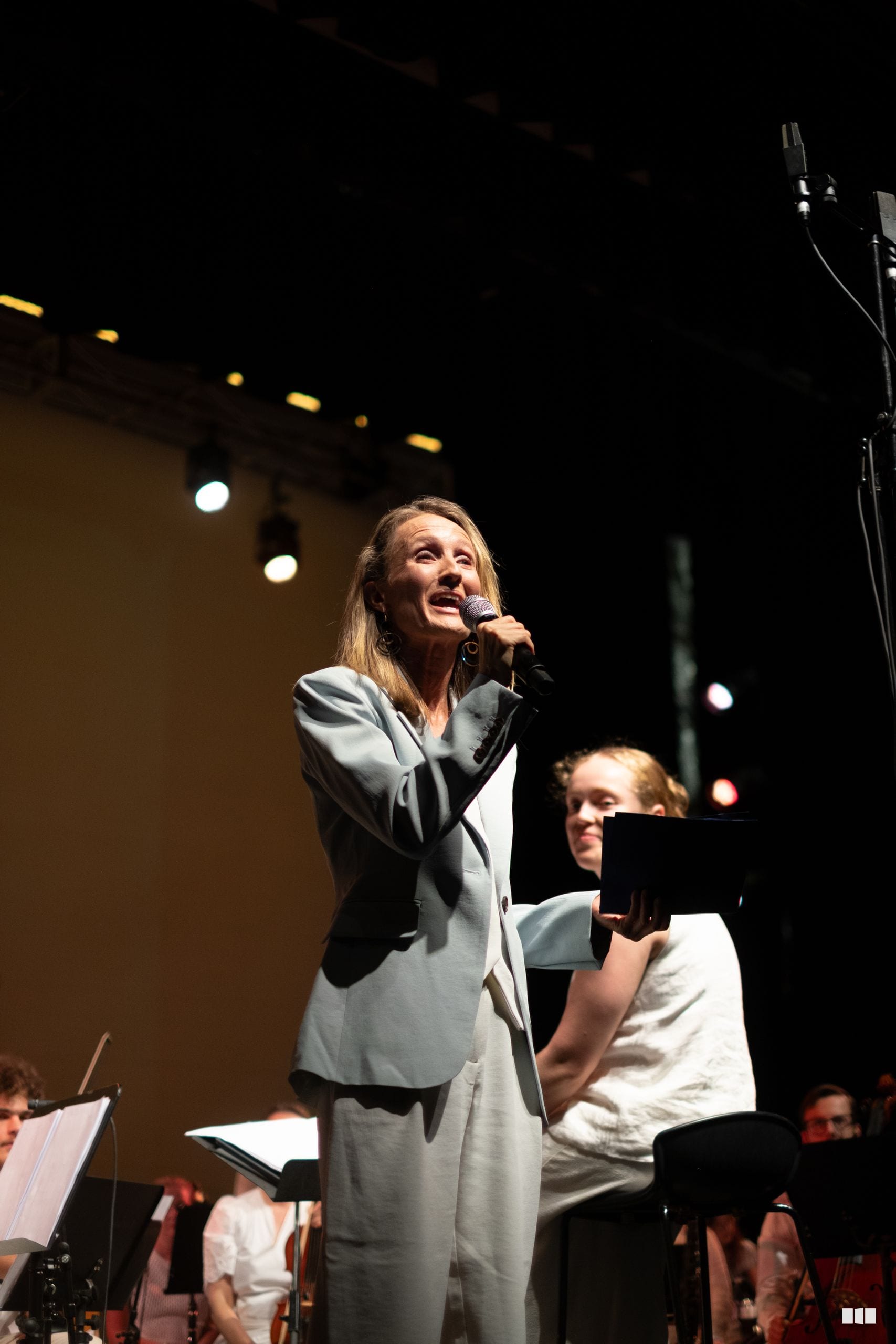 A woman in a light grey blazer sings into a microphone on stage while a young pianist accompanies her during a performance.