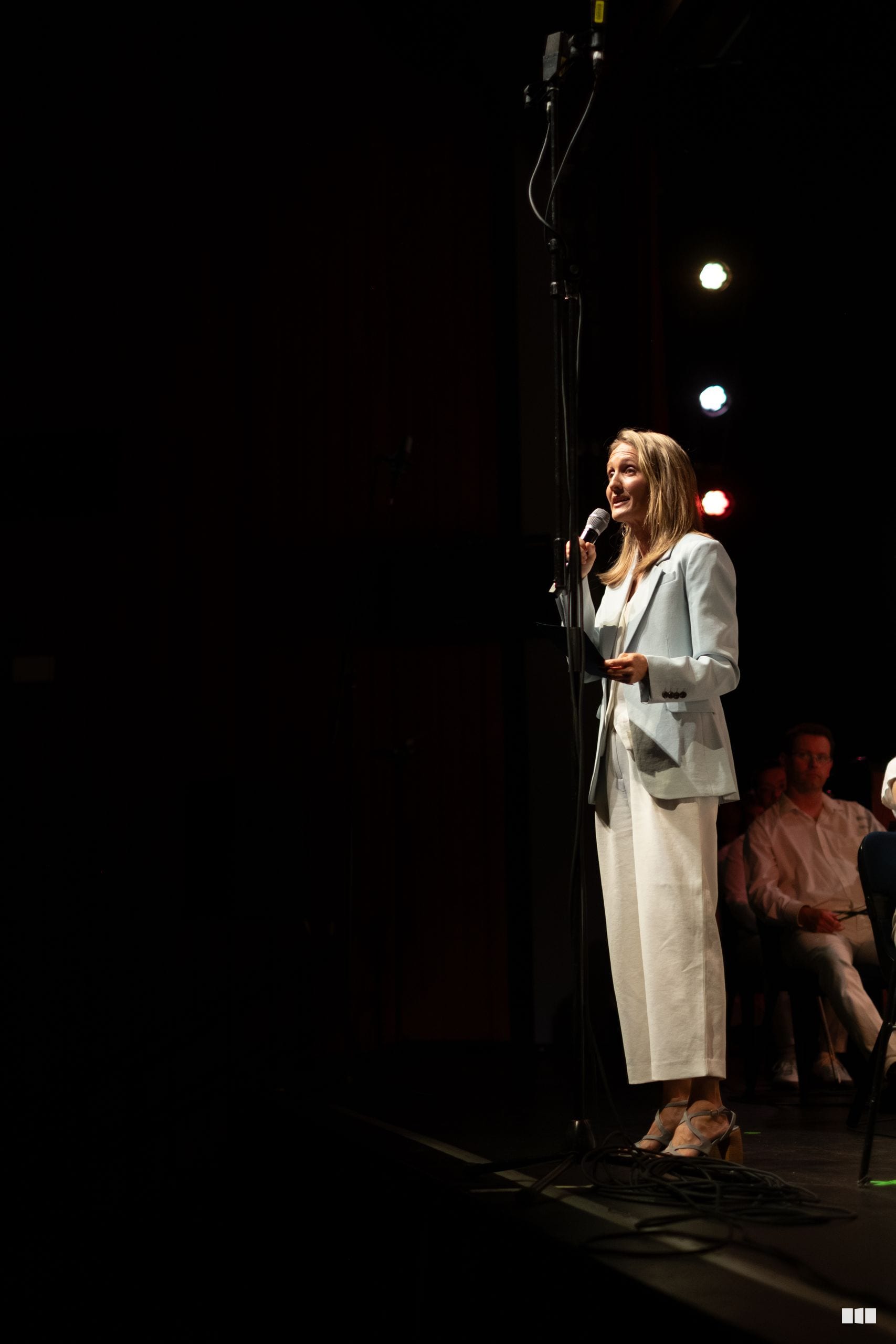 A woman in a cream blazer speaks at a microphone on stage under theatrical lighting.