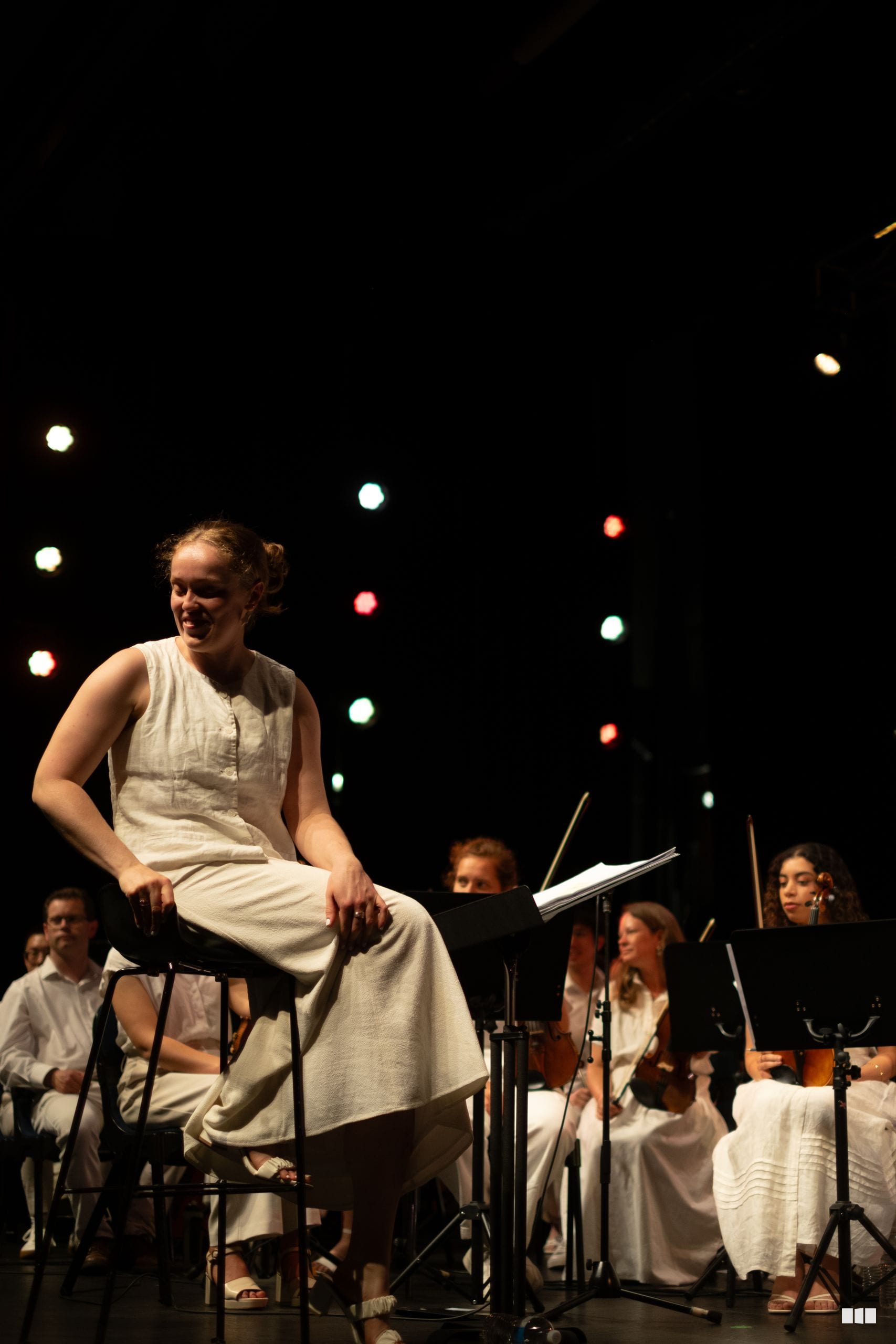 A young female soloist performs on stage with orchestra members seated behind her against a dark backdrop with stage lighting.