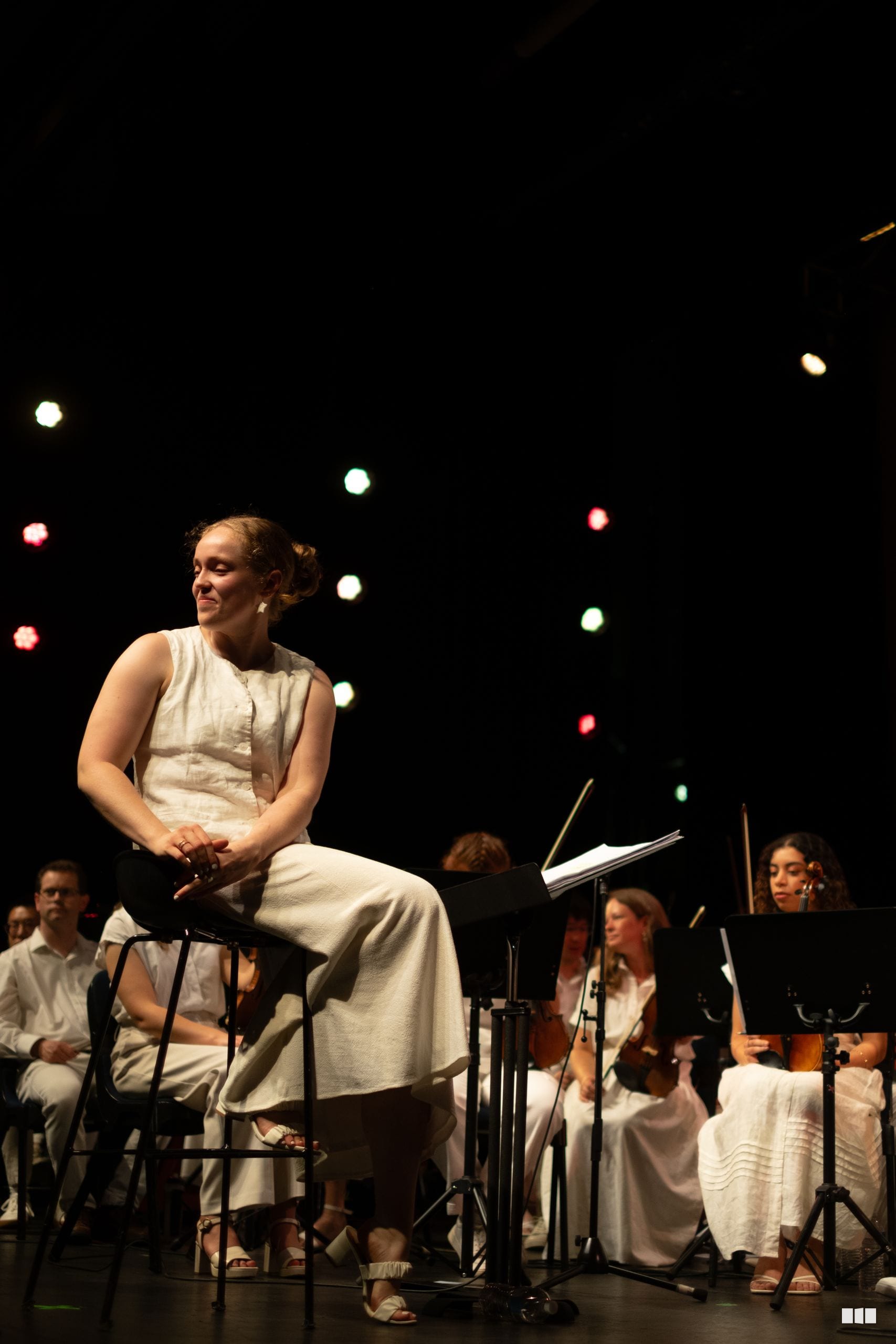 A soloist in a cream-colored dress sits on a stool before an ensemble of string and wind musicians on stage with atmospheric lighting.