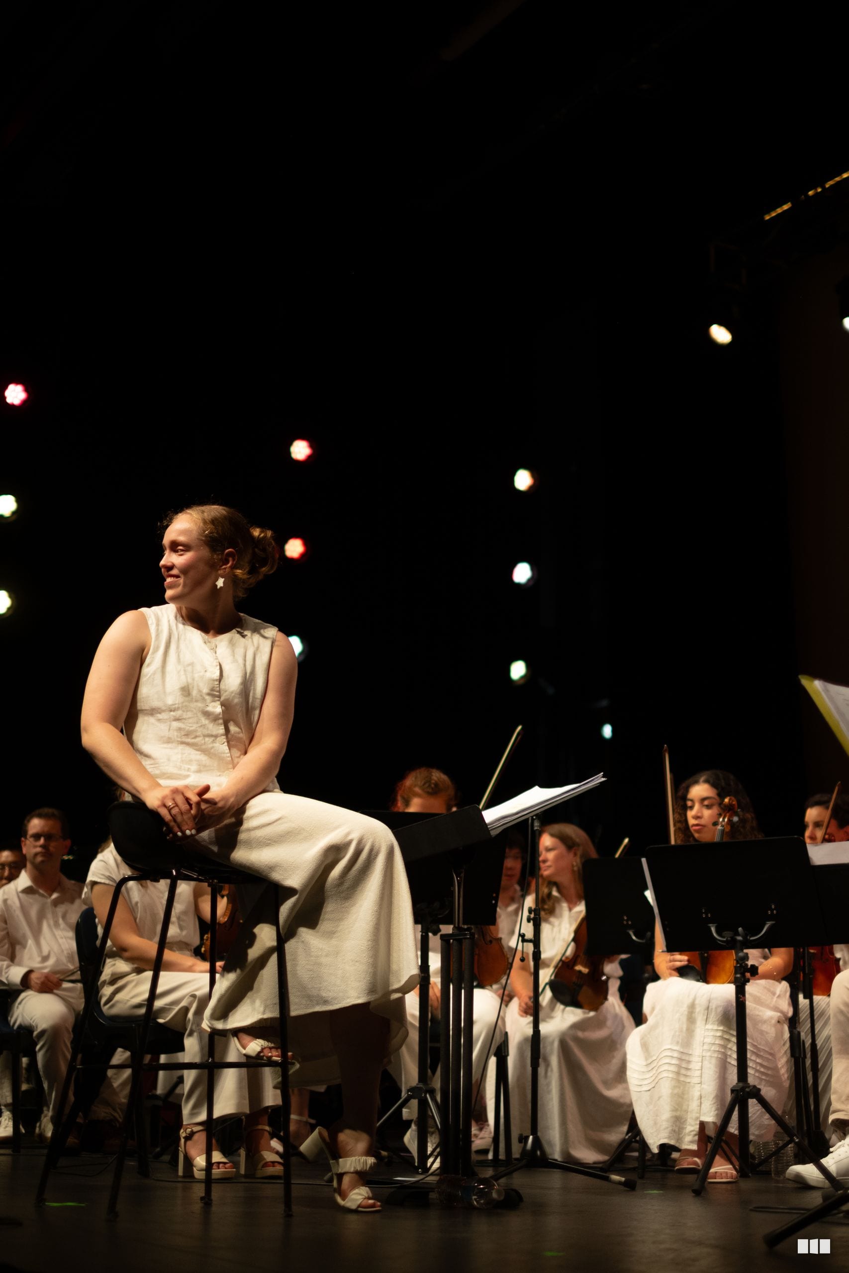 A female soloist sits on stage surrounded by orchestra members during a performance against a backdrop of warm stage lighting.