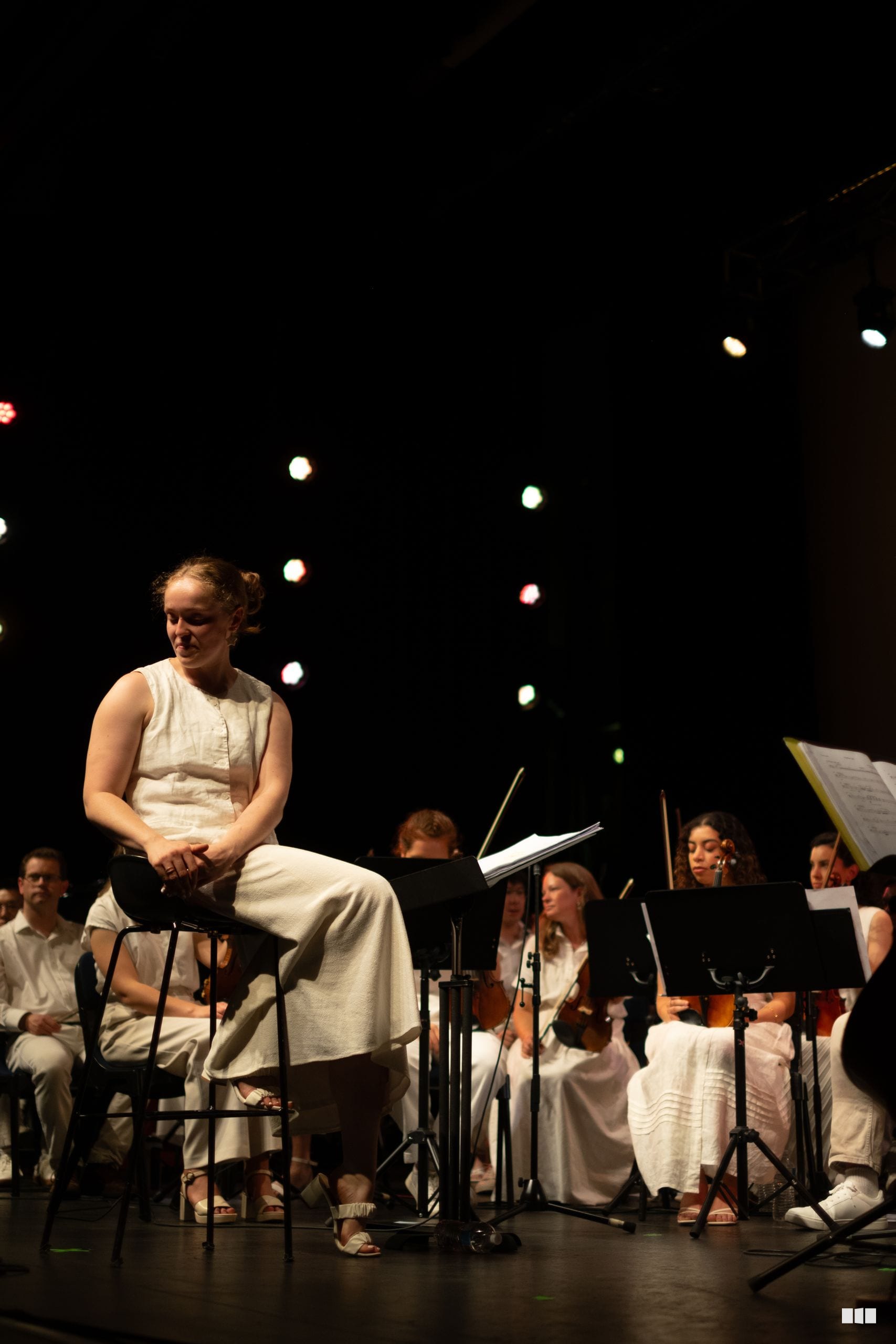 A soloist sits thoughtfully on stage surrounded by orchestra members during a performance under stage lighting.