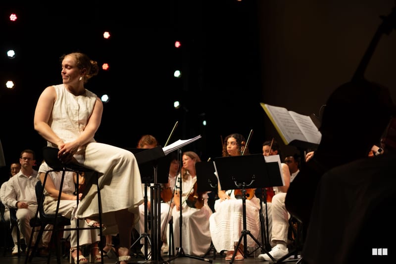 A soloist in a cream-colored dress performs with an orchestra during a concert, with atmospheric stage lighting in the background.