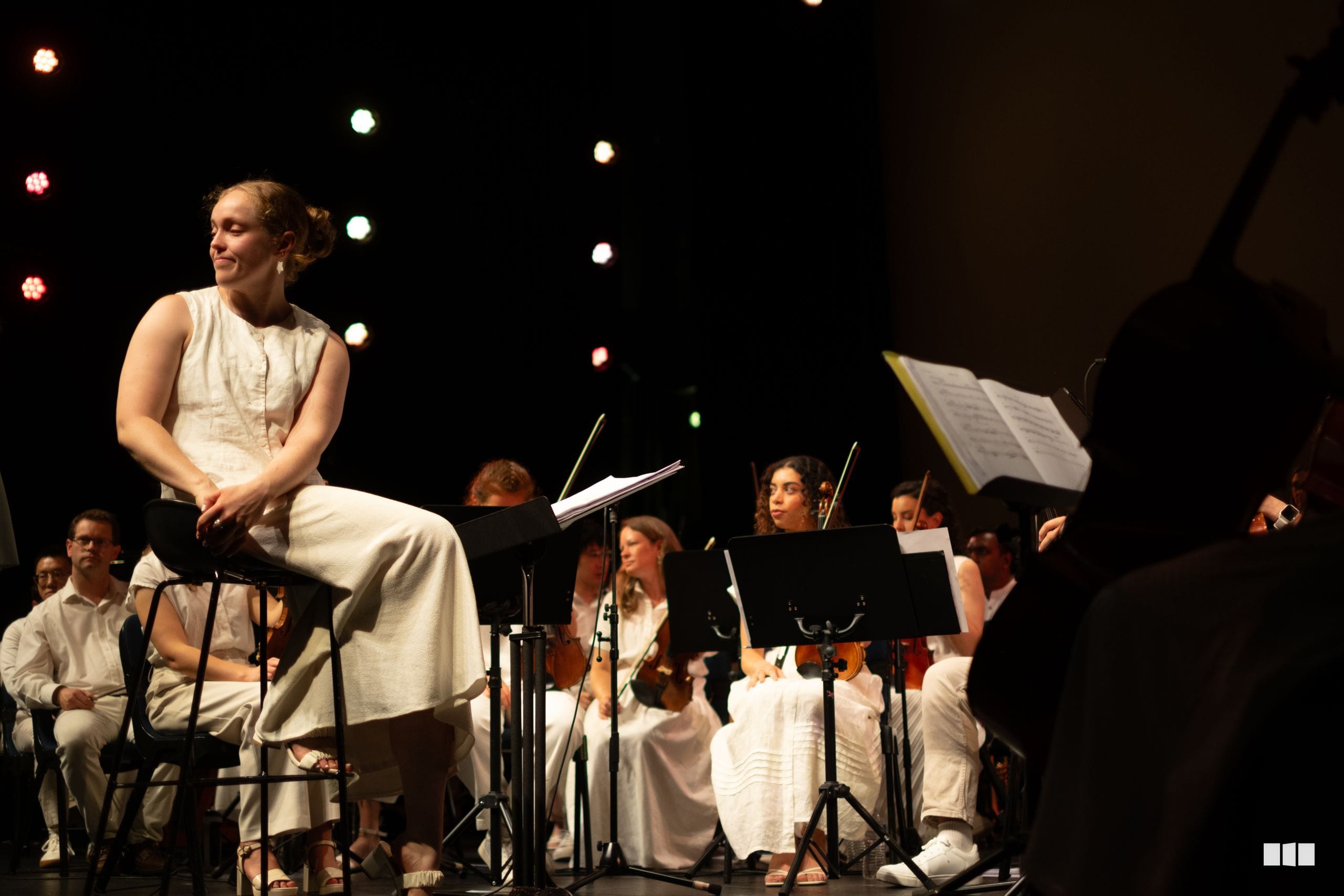 A soloist in a cream-colored dress sits on stage with orchestra members in white attire, accompanied by sheet music stands and warm stage lighting.