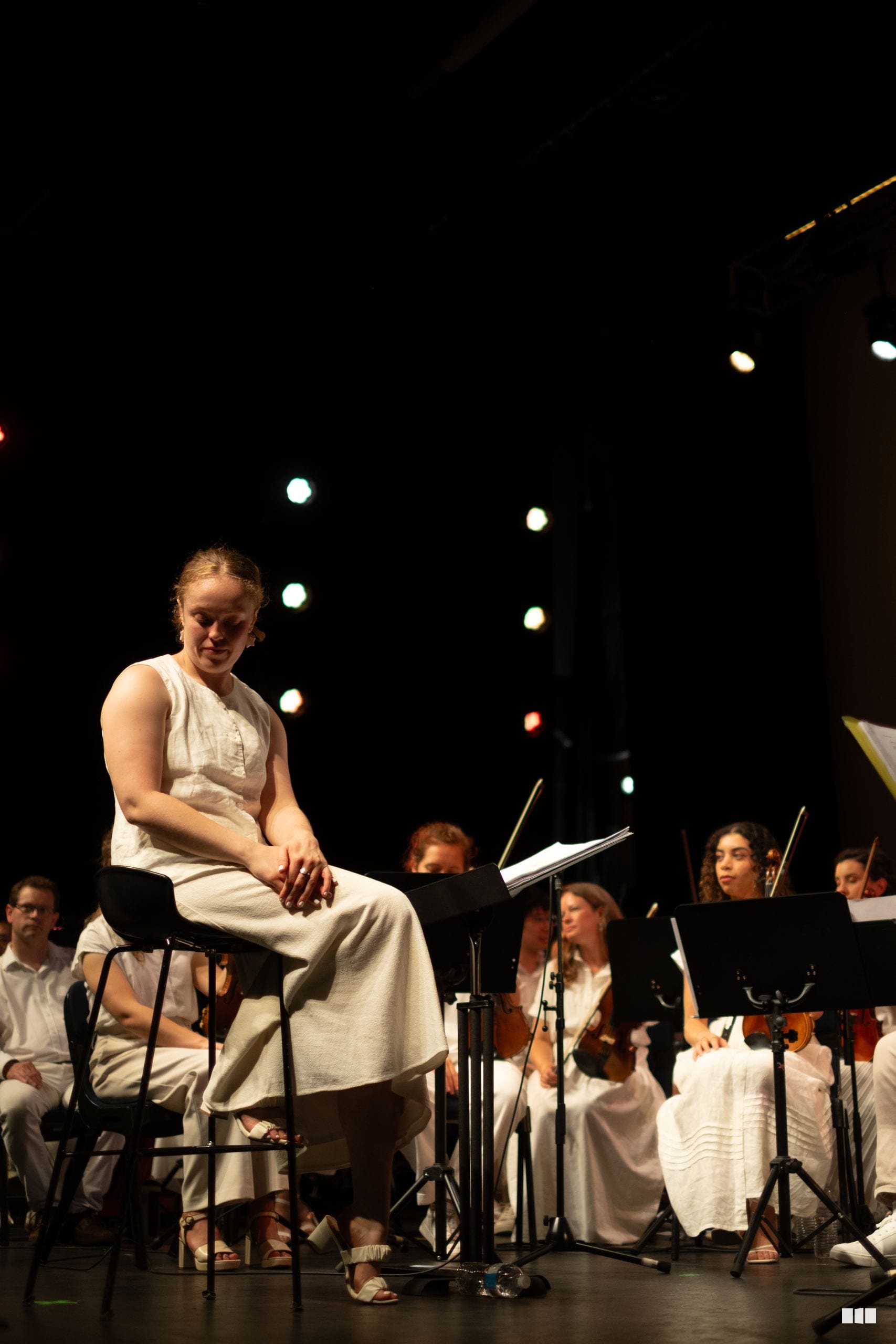 A soloist in a light-colored dress performs with an orchestra ensemble on stage during an evening concert.